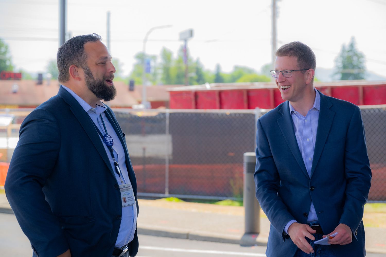 Two men in dark blazers stand outdoors talking near a fence and red barriers, one bearded with a lanyard, the other wearing glasses.