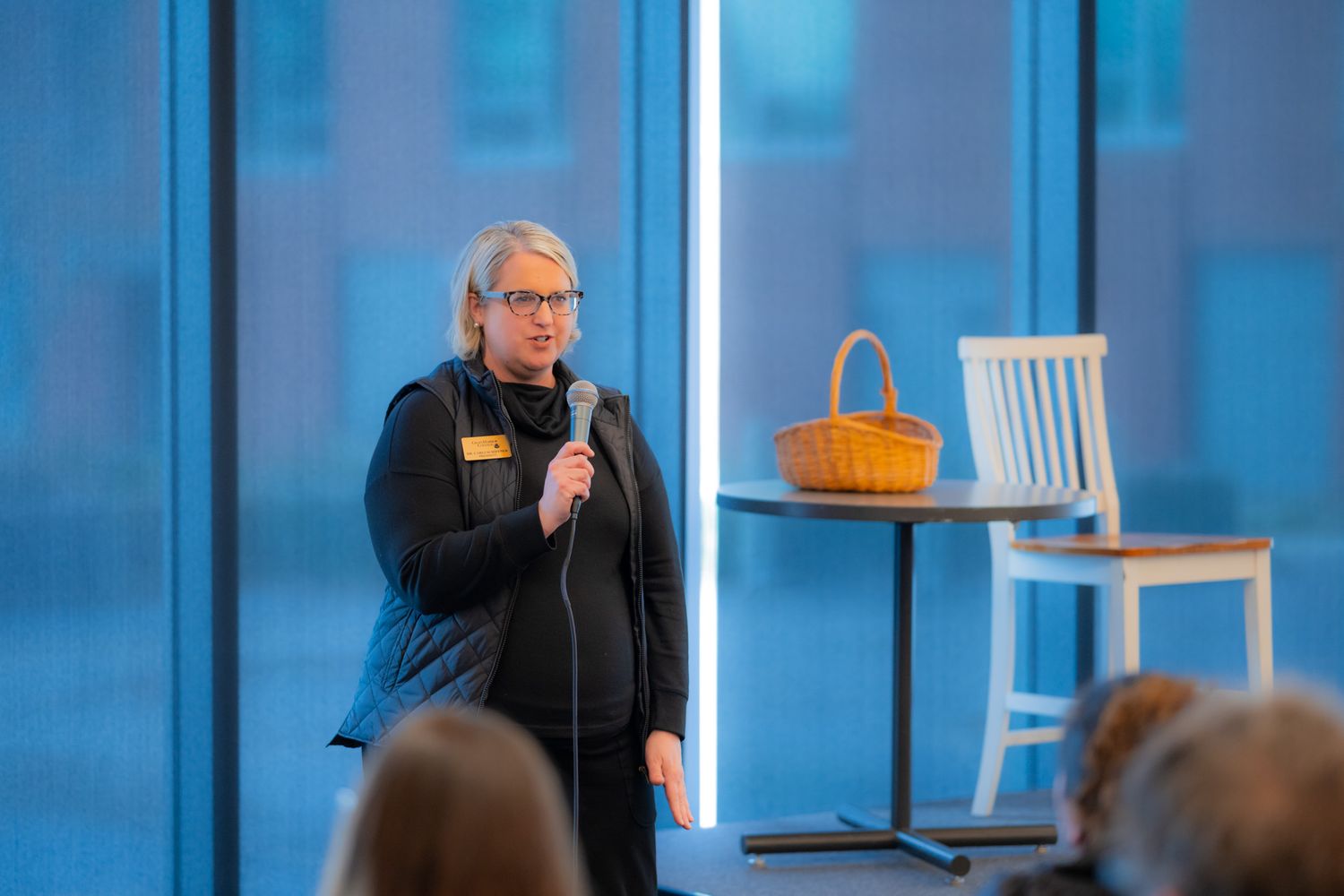 A woman with glasses and a name tag speaks into a microphone to a seated audience near large windows, with a wicker basket on a table.