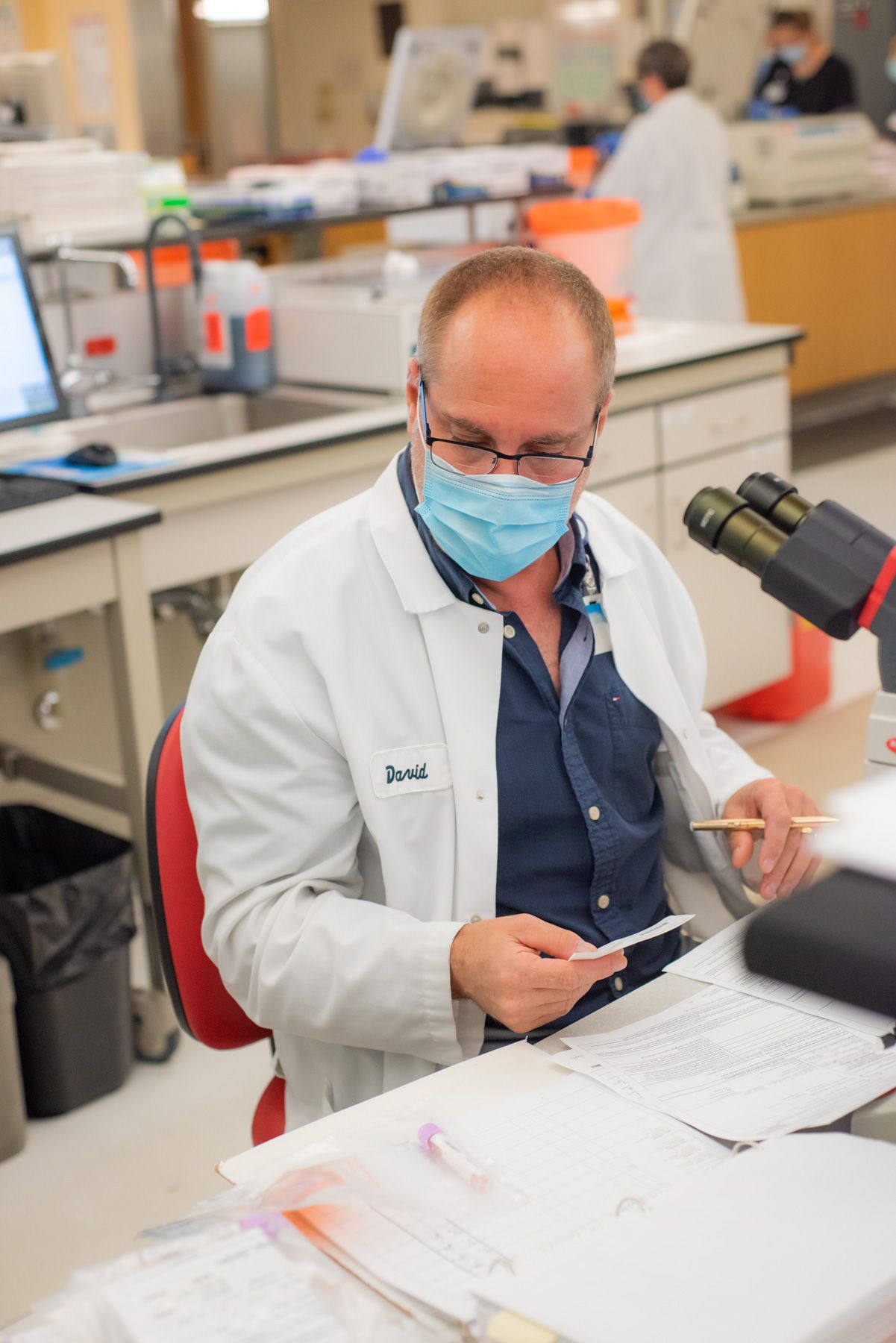 Laboratory technician in a white coat and surgical mask reviews paperwork at a lab workstation with a microscope nearby.