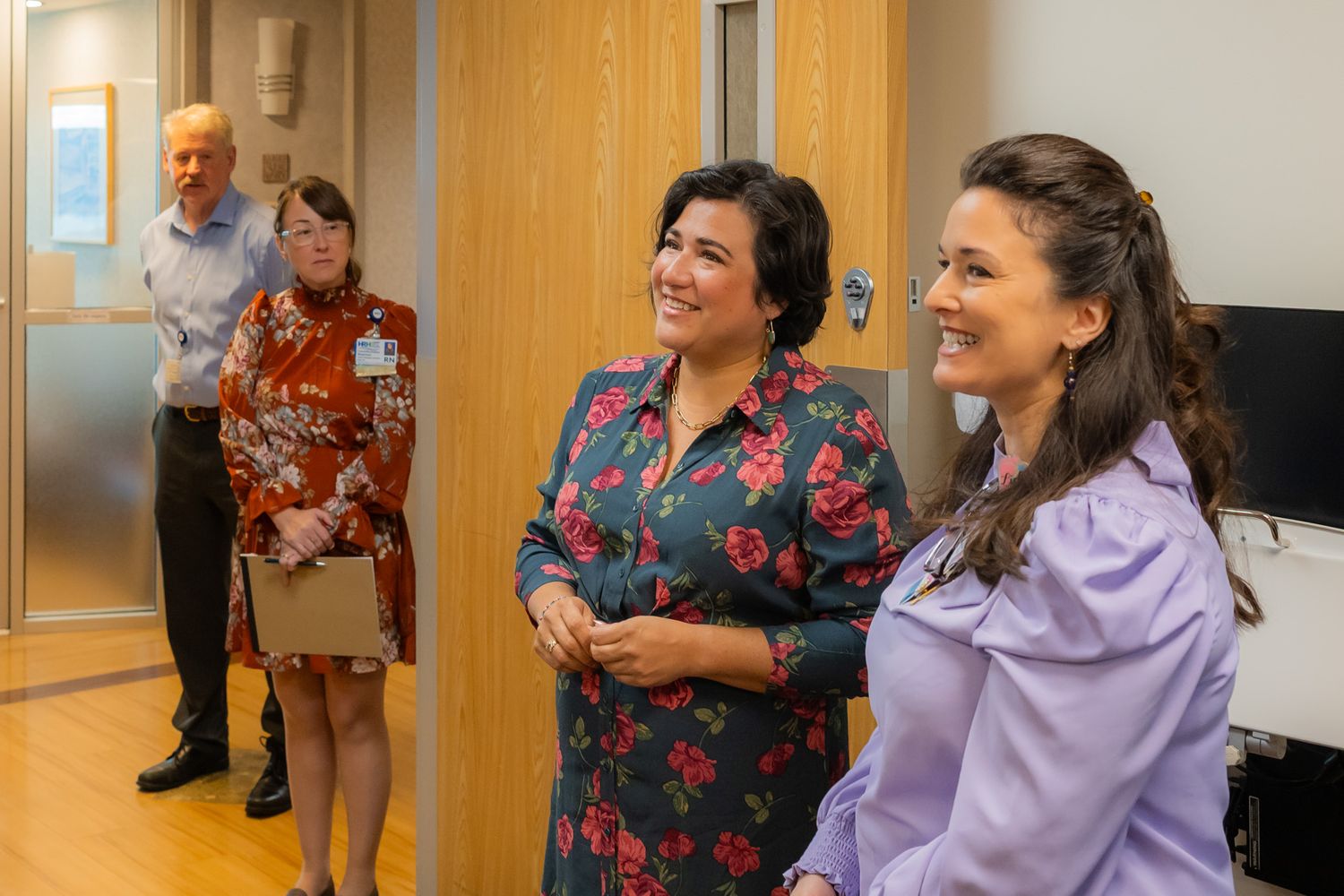 Two smiling women in floral and lavender blouses stand in a bright hallway while a man and woman talk in the background.