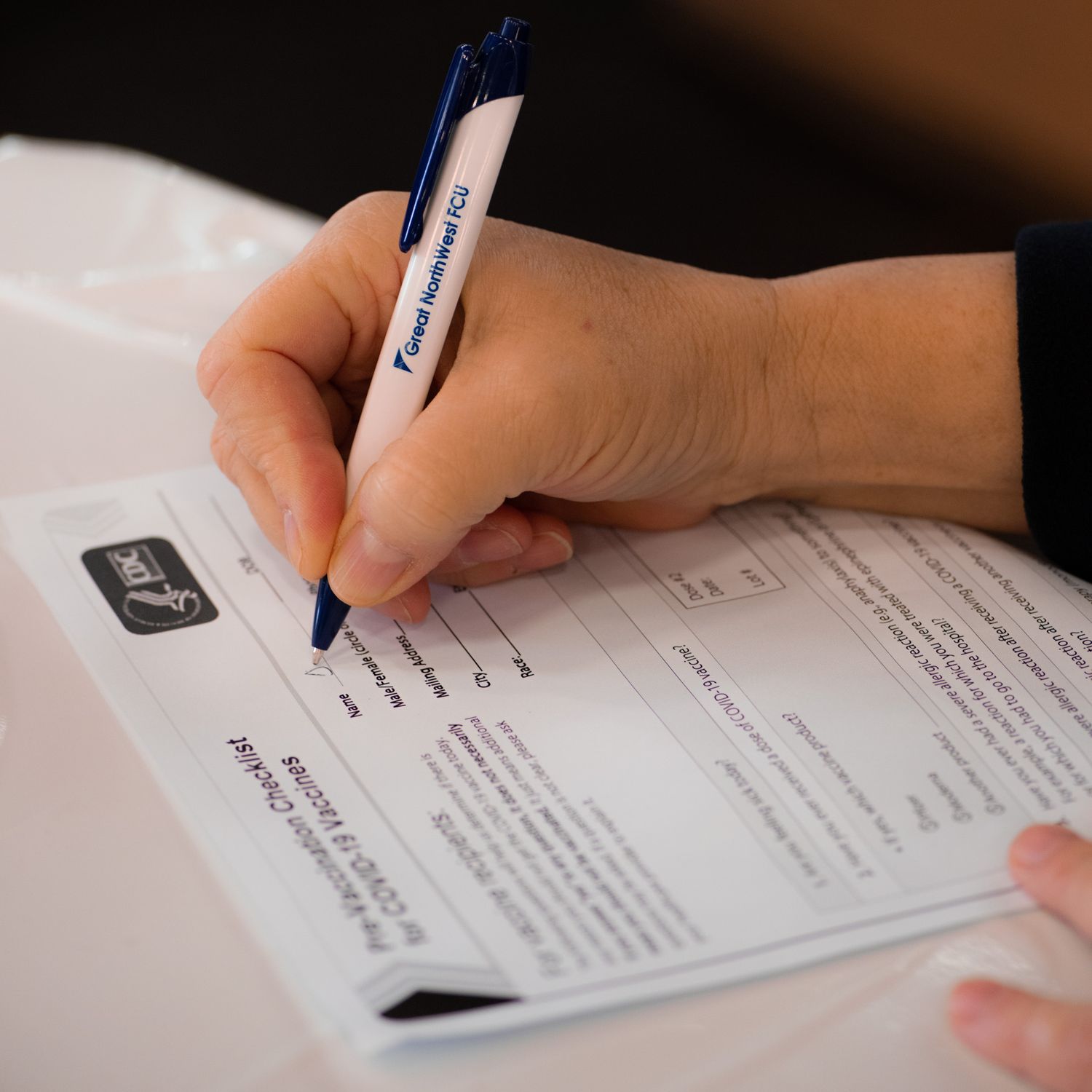 A person fills out a COVID-19 vaccine consent form on a table using a pen, with the paperwork resting on a white tablecloth.