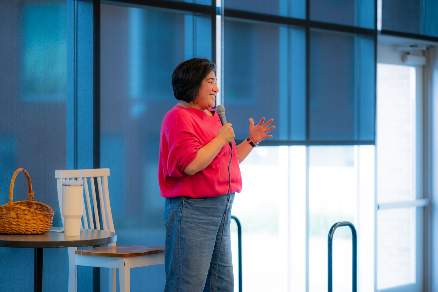 Woman in a bright pink top and jeans speaks into a microphone while gesturing on stage beside a wicker basket and chair near large windows.