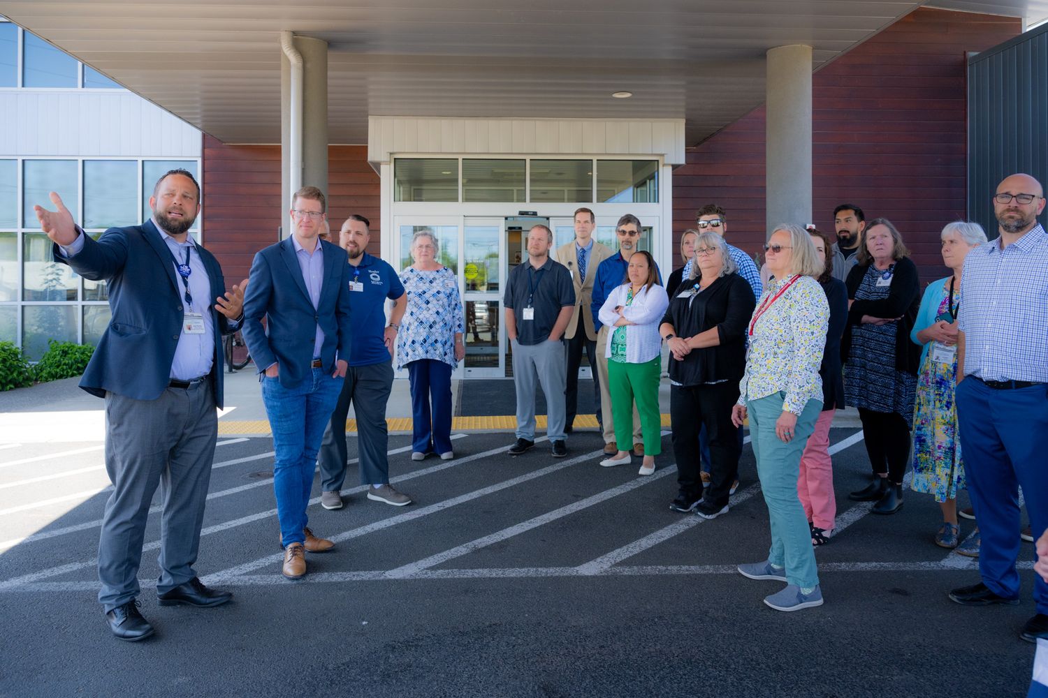 Group of people gathered outside a brick building entrance while a man in a blazer gestures and speaks to the crowd.