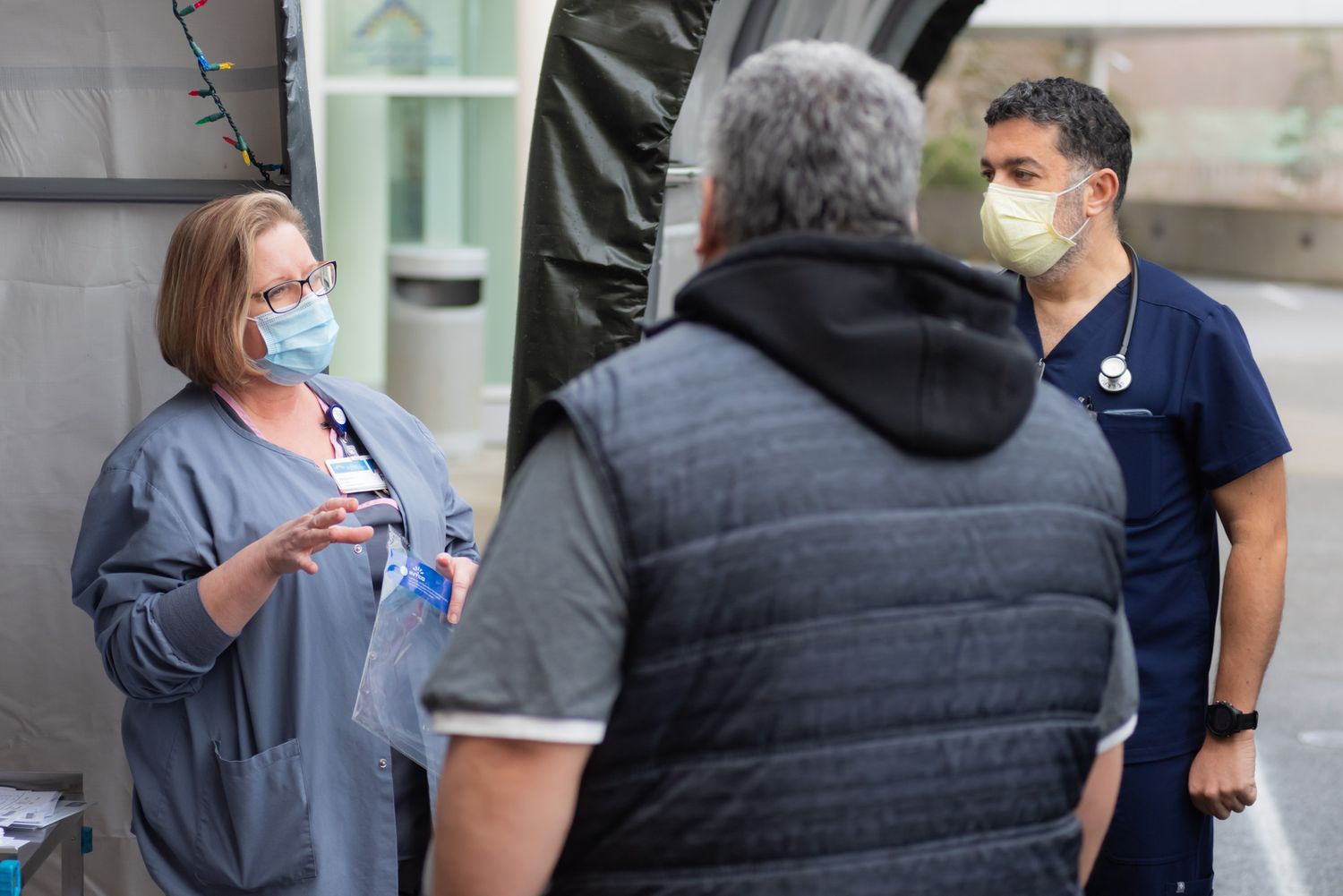 Two healthcare workers in scrubs and face masks talk with a man in a gray vest outside a medical tent at an outdoor care site.