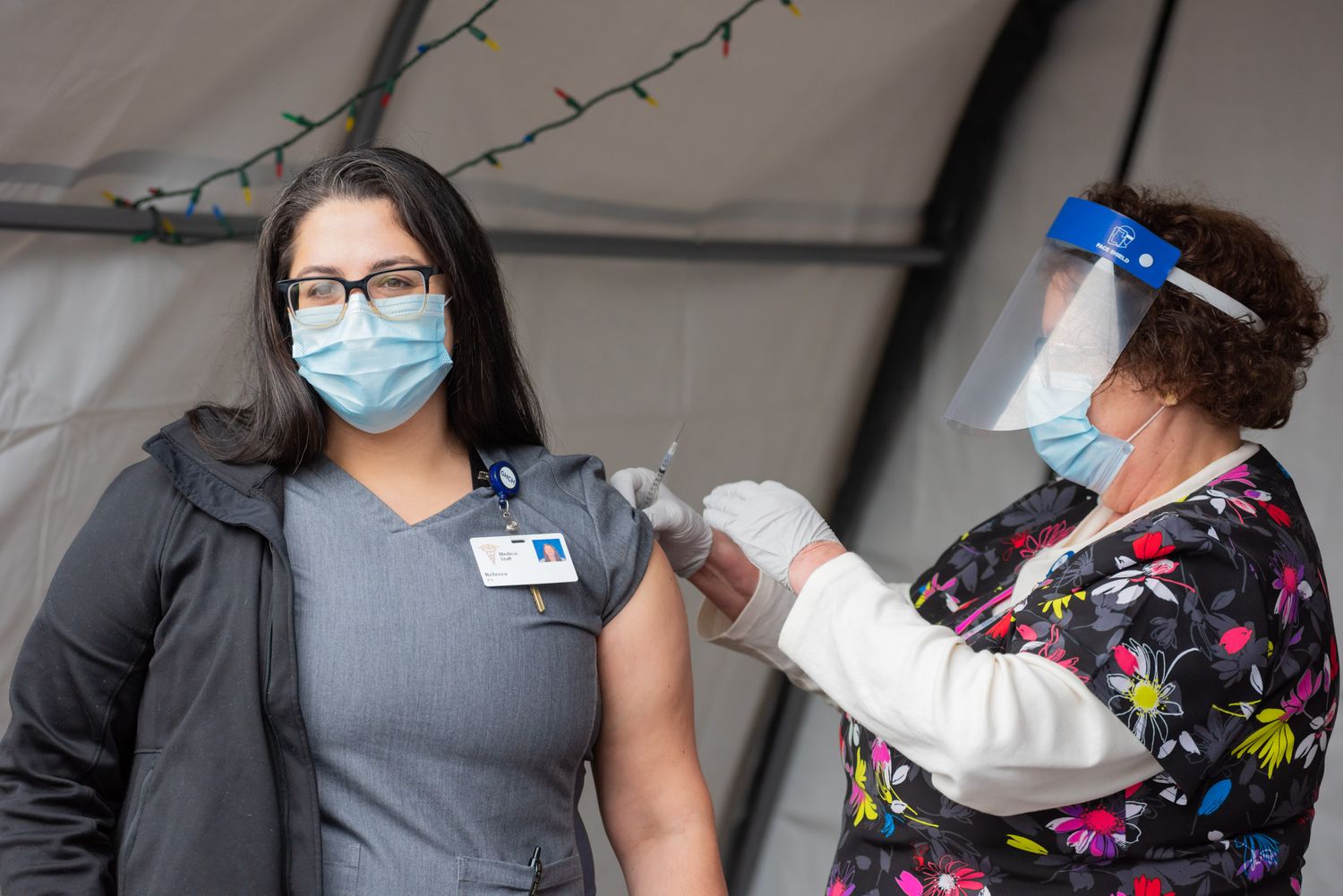 Healthcare worker in a face shield and floral scrubs gives a vaccine injection to a masked woman in gray scrubs at an outdoor clinic tent.