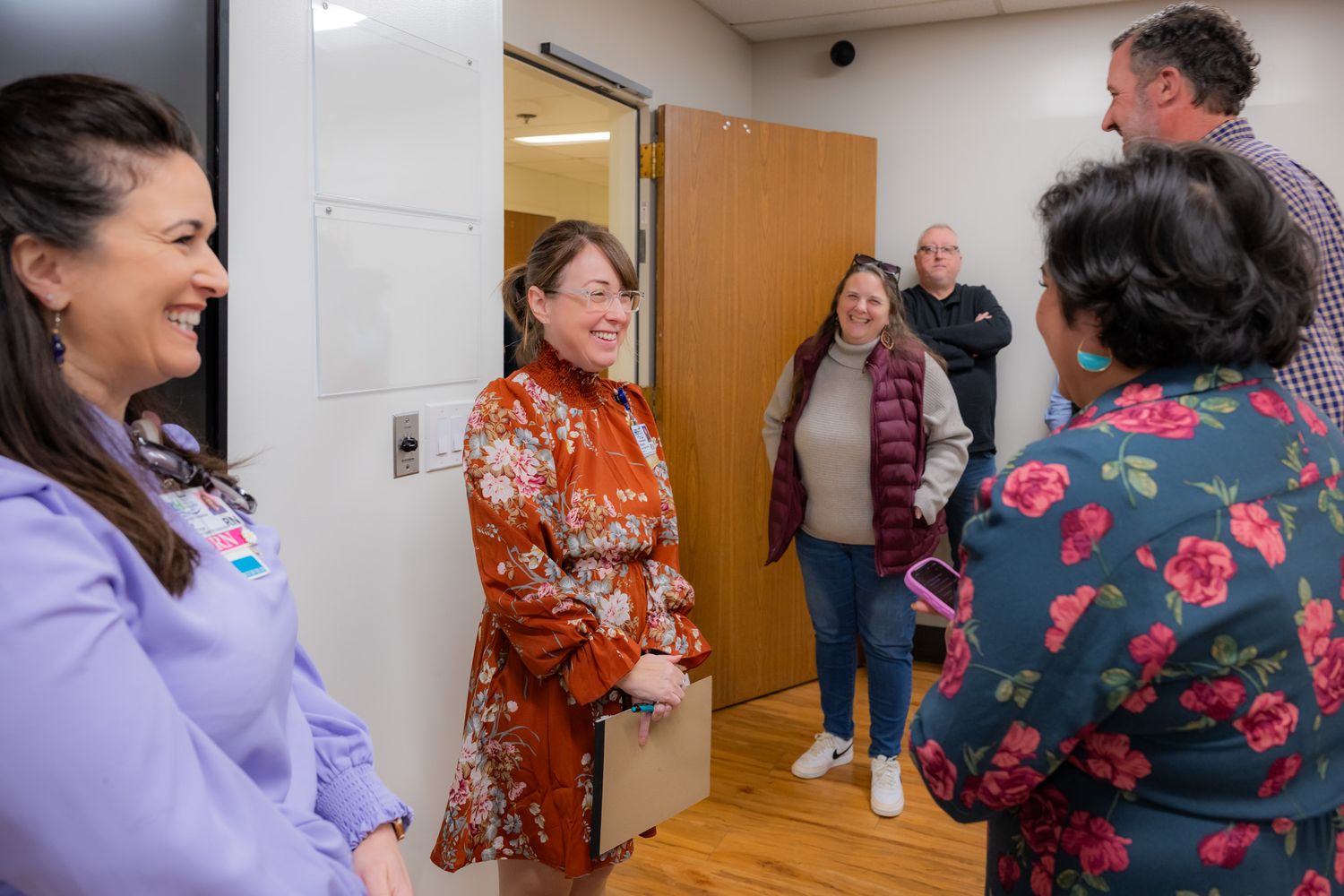 A group of smiling women and men chat in a bright office hallway, with one woman in a floral blouse holding a folder near an open door.