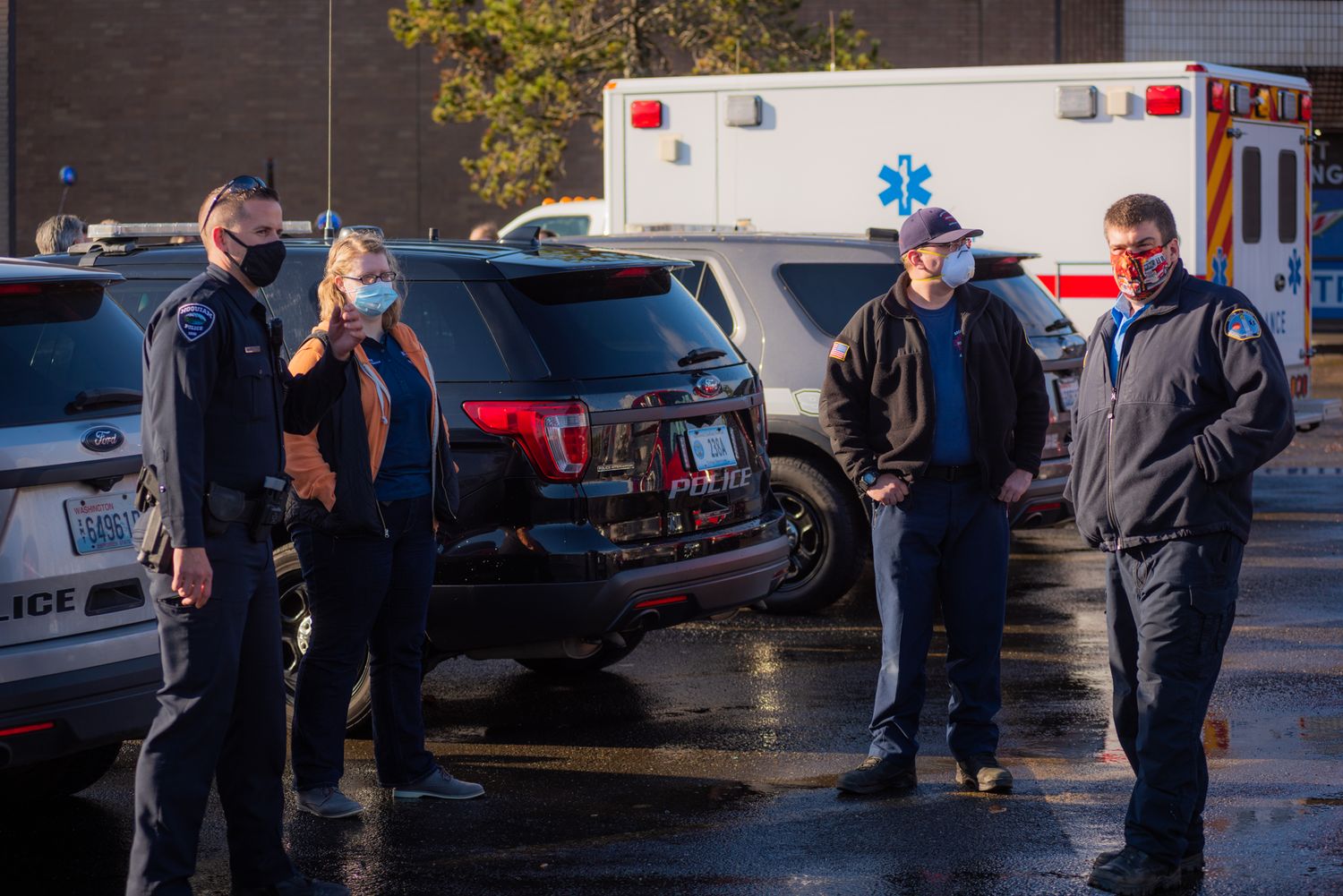 Masked police officers and emergency responders stand near patrol SUVs and an ambulance in a wet parking lot on an overcast day.