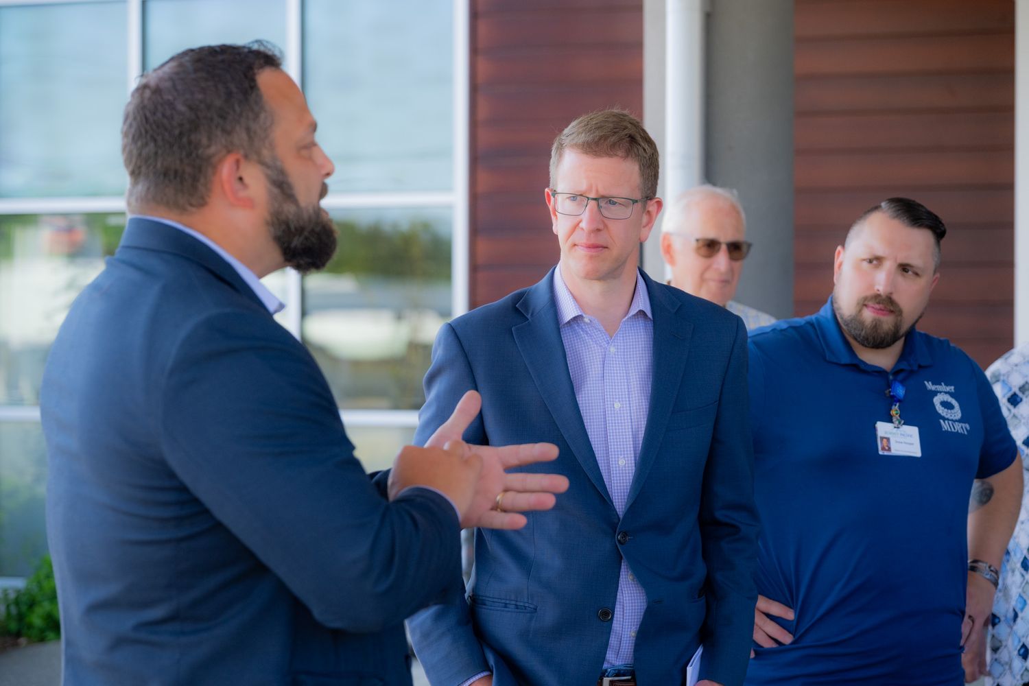 A bearded man in a navy blazer gestures while speaking to a group of men in business attire outside a modern building.