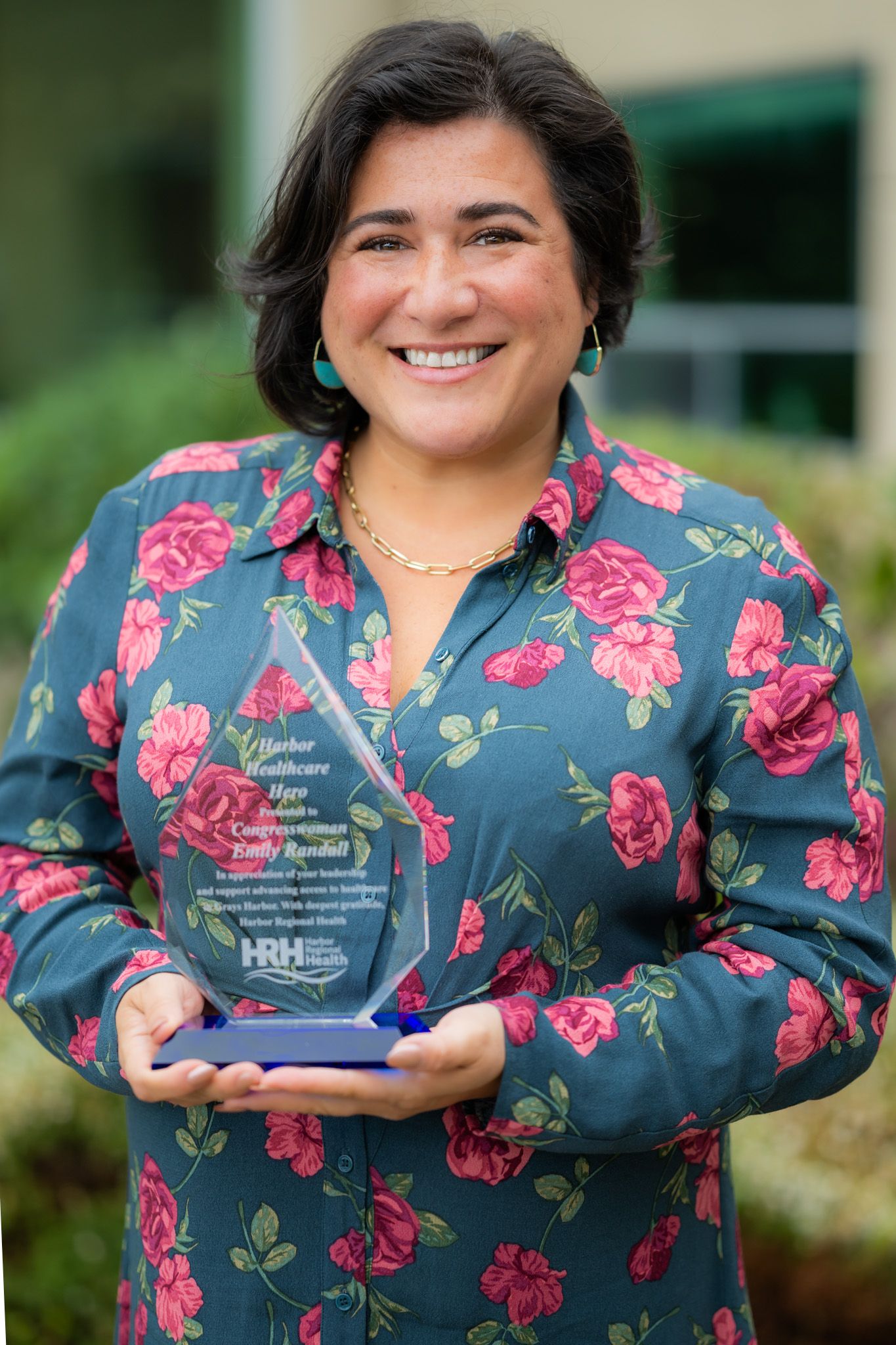 Smiling woman in a floral teal blouse holds a glass recognition award, standing outdoors in front of green shrubs and a building.