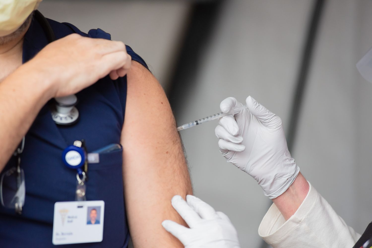 Healthcare worker in navy scrubs and a face mask receives a vaccine injection in the upper arm from a gloved clinician.