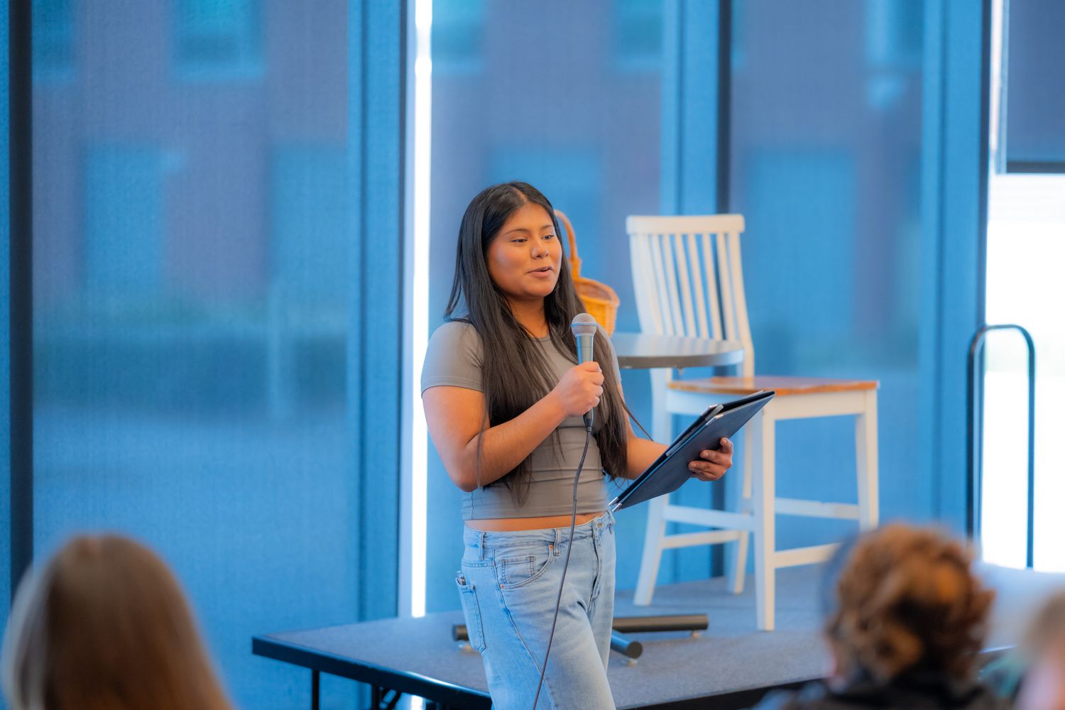 A woman with long dark hair speaks into a microphone while holding a tablet, standing before a seated audience near large windows.