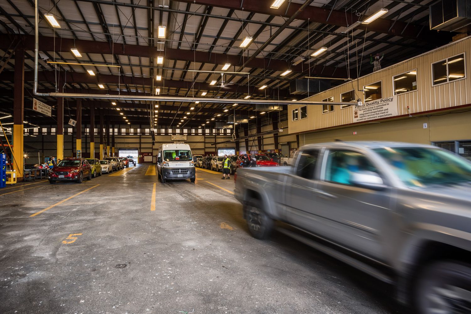 A silver pickup truck drives through a large covered warehouse lot as vehicles and a white van are parked along the walls.