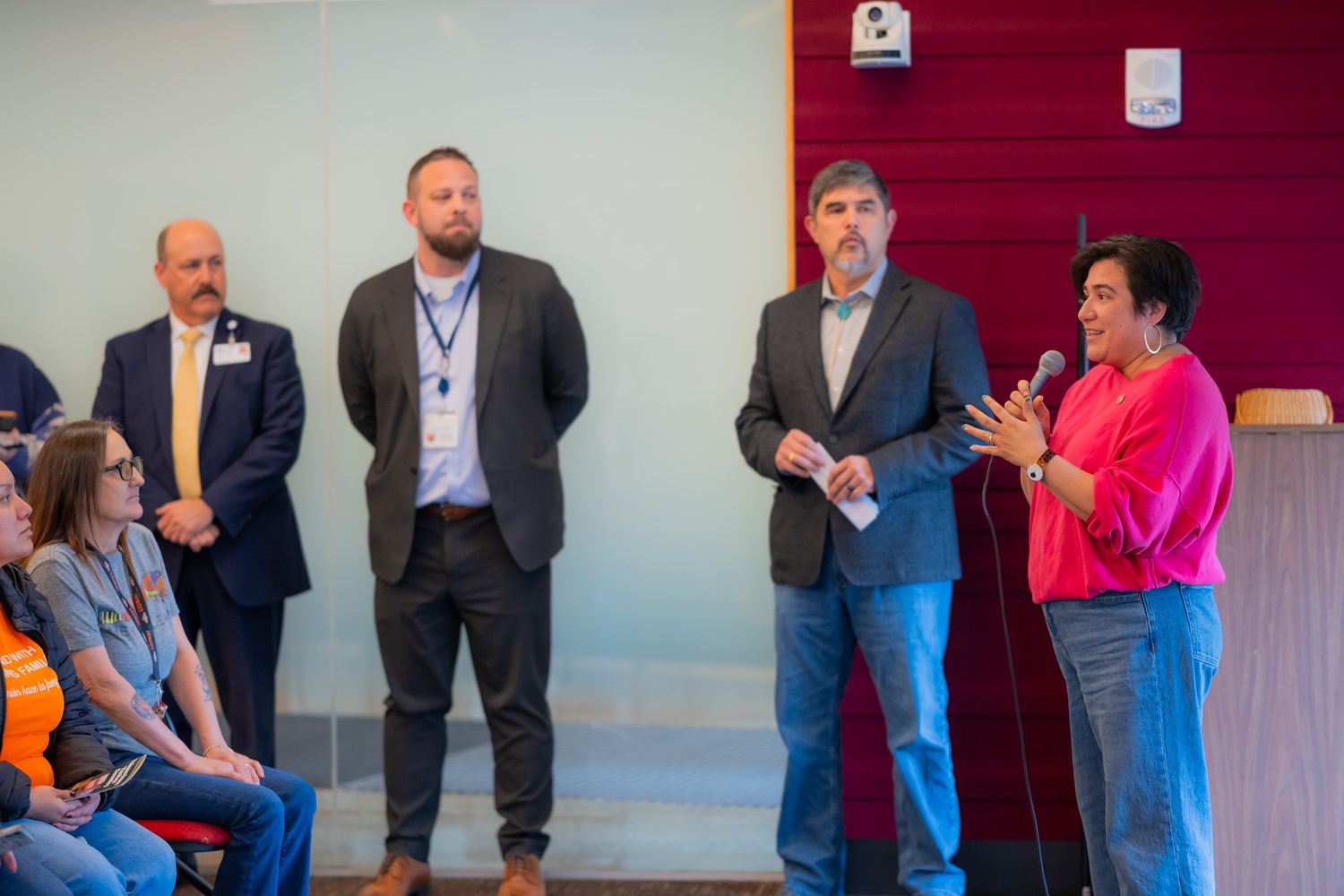 A woman in a pink top speaks into a microphone as three men in blazers stand beside her at a community event.