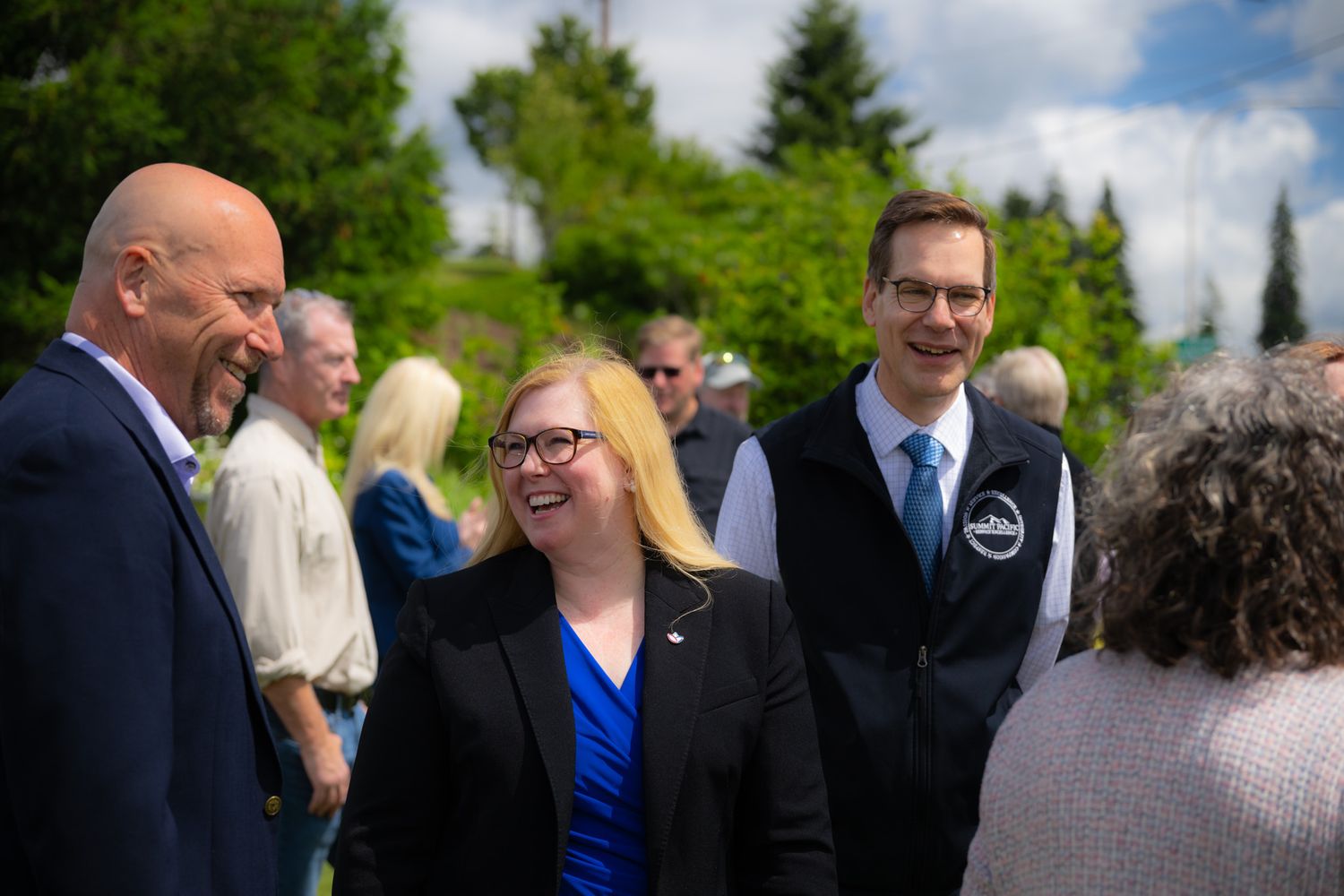 Smiling woman in a black blazer and glasses chats with a man in a vest and tie at a sunny outdoor gathering surrounded by trees.