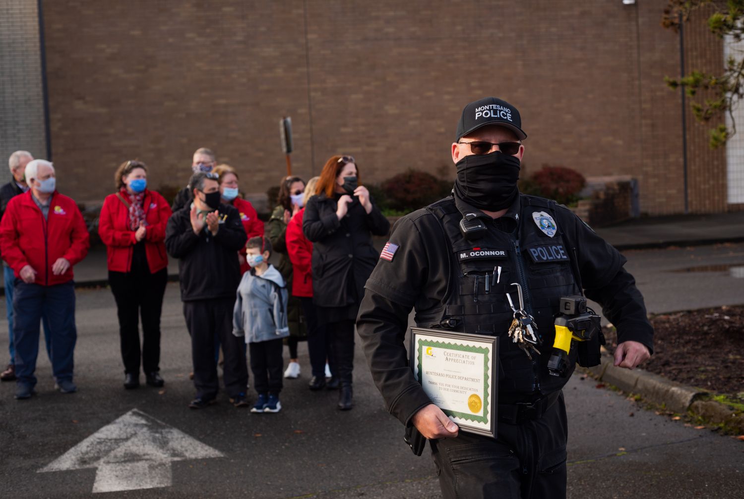 A masked police officer carries a framed certificate while smiling onlookers in red jackets watch in the background.