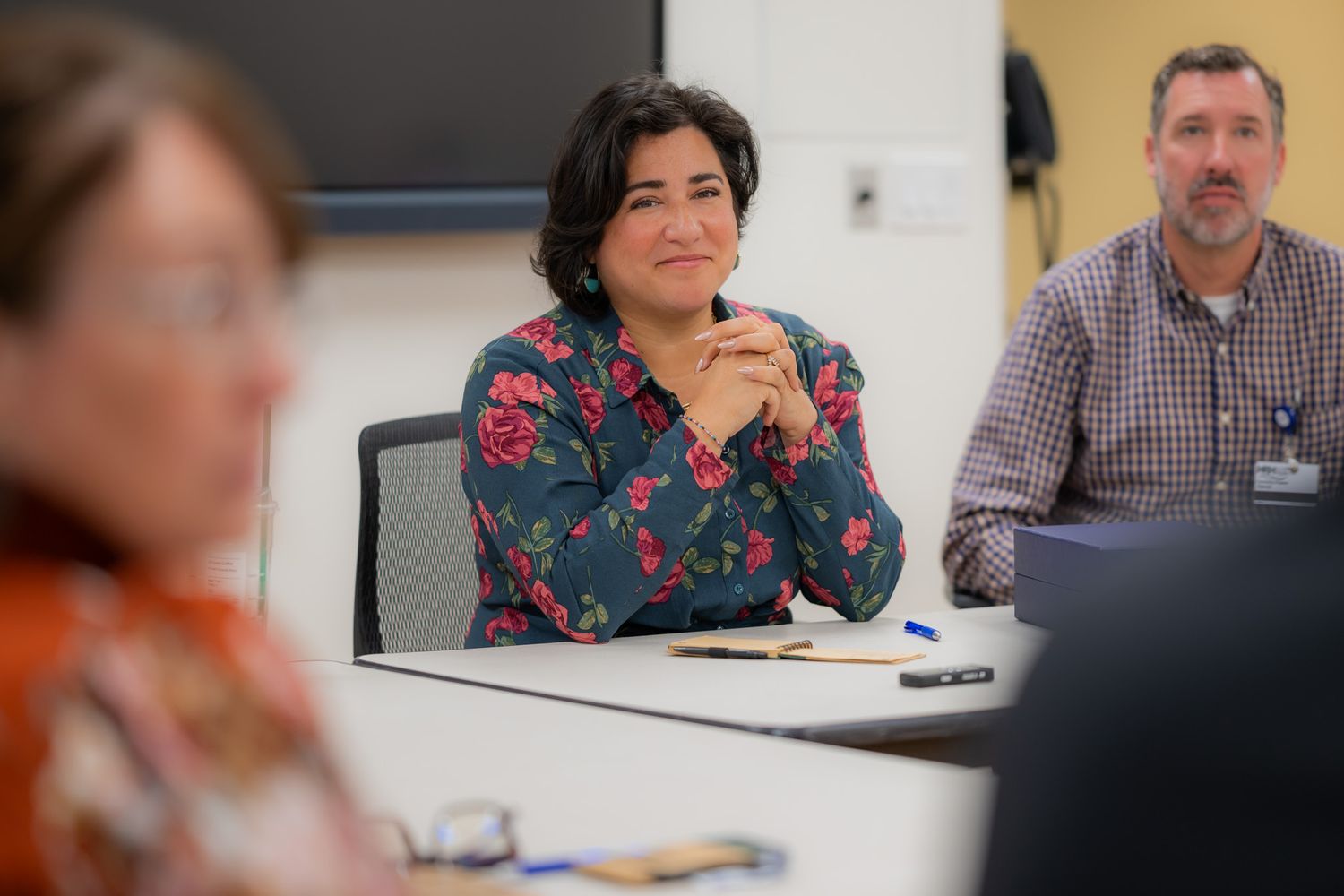 Smiling woman in a floral blouse sits with clasped hands at a conference table while colleagues listen during a meeting.