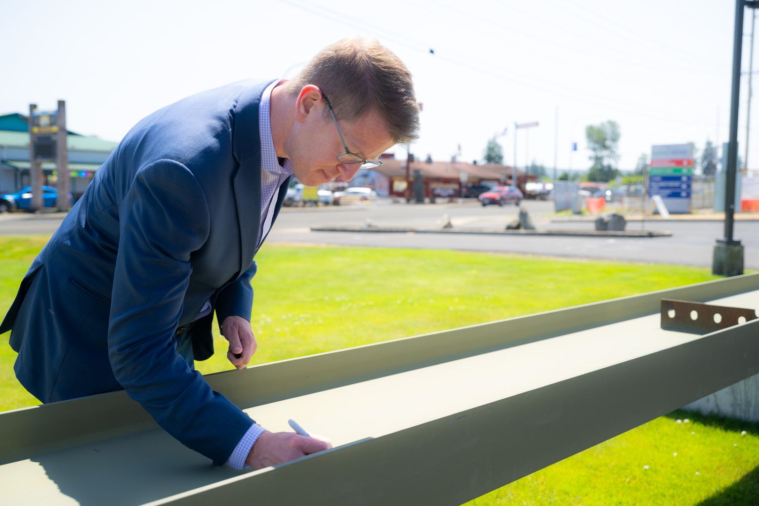 Man in a navy suit signs a green steel beam outdoors on a sunny day, with a grassy lawn and road in the background.