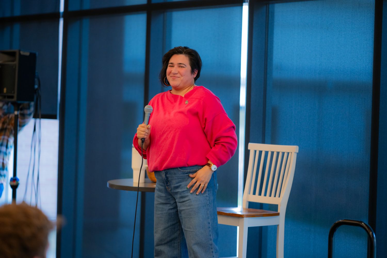 A smiling woman in a pink sweater and jeans stands with a microphone next to a white chair and a small round table.