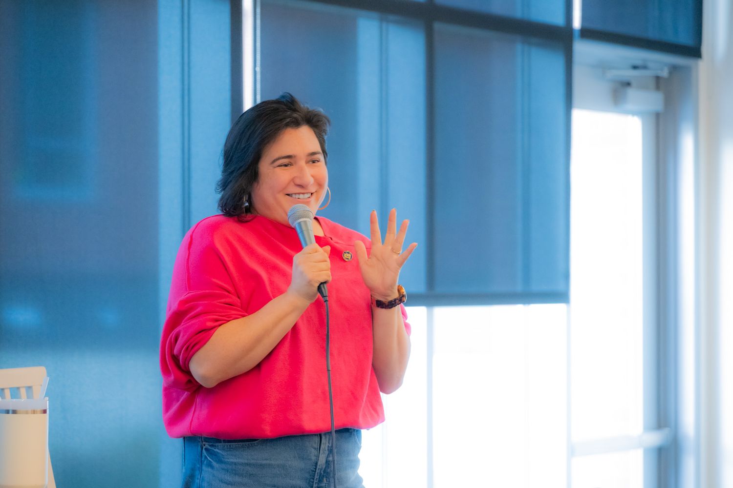 A smiling woman in a bright pink top gestures with one hand while holding a microphone during a speaking engagement.