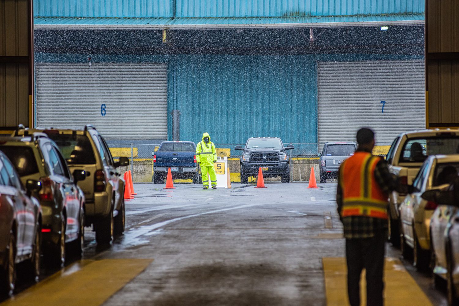 Staff in high-visibility gear guide lines of cars past orange traffic cones at a drive-through site outside a large warehouse.