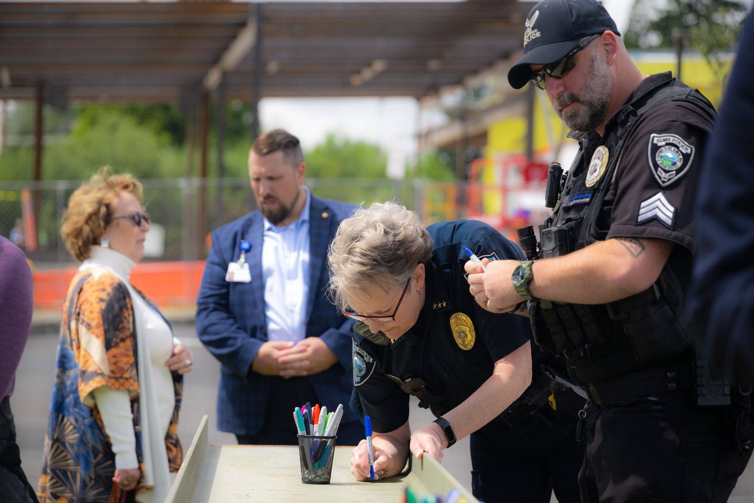 Uniformed police officers sign paperwork at an outdoor table while a woman in a colorful cardigan and a man in a blue blazer look on.
