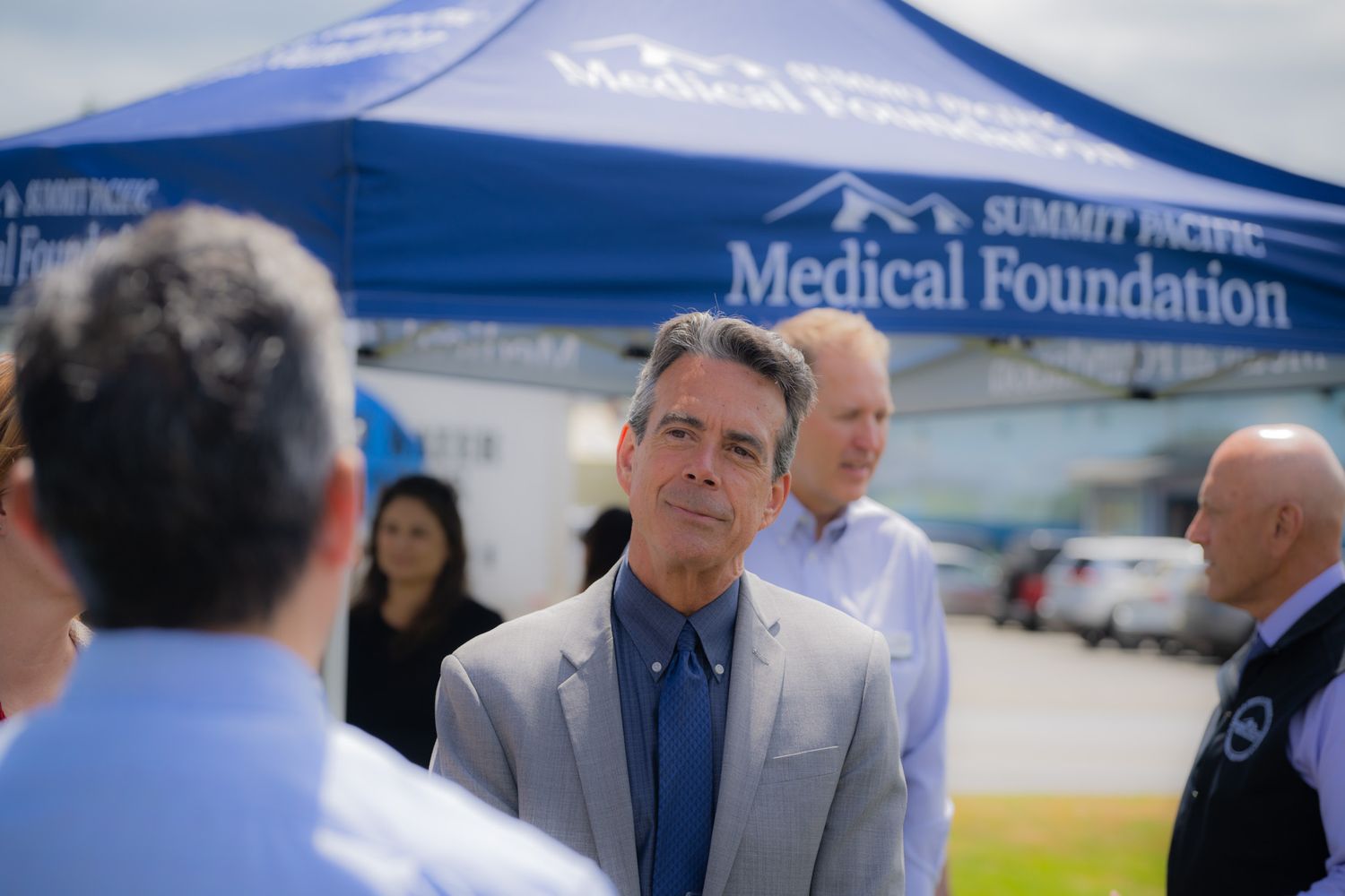 Man in a gray blazer stands outdoors near a blue Summit Pacific Medical Foundation tent as people gather in the background.
