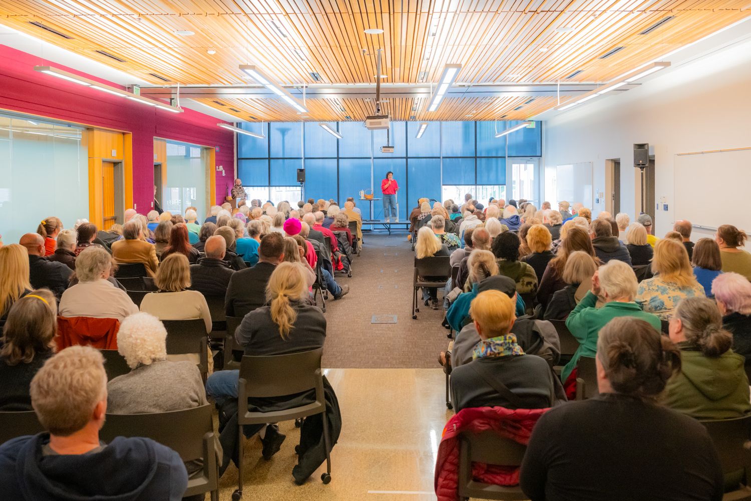Large audience seated in rows listening to a speaker at the front of a bright community hall with wood ceiling and tall windows.