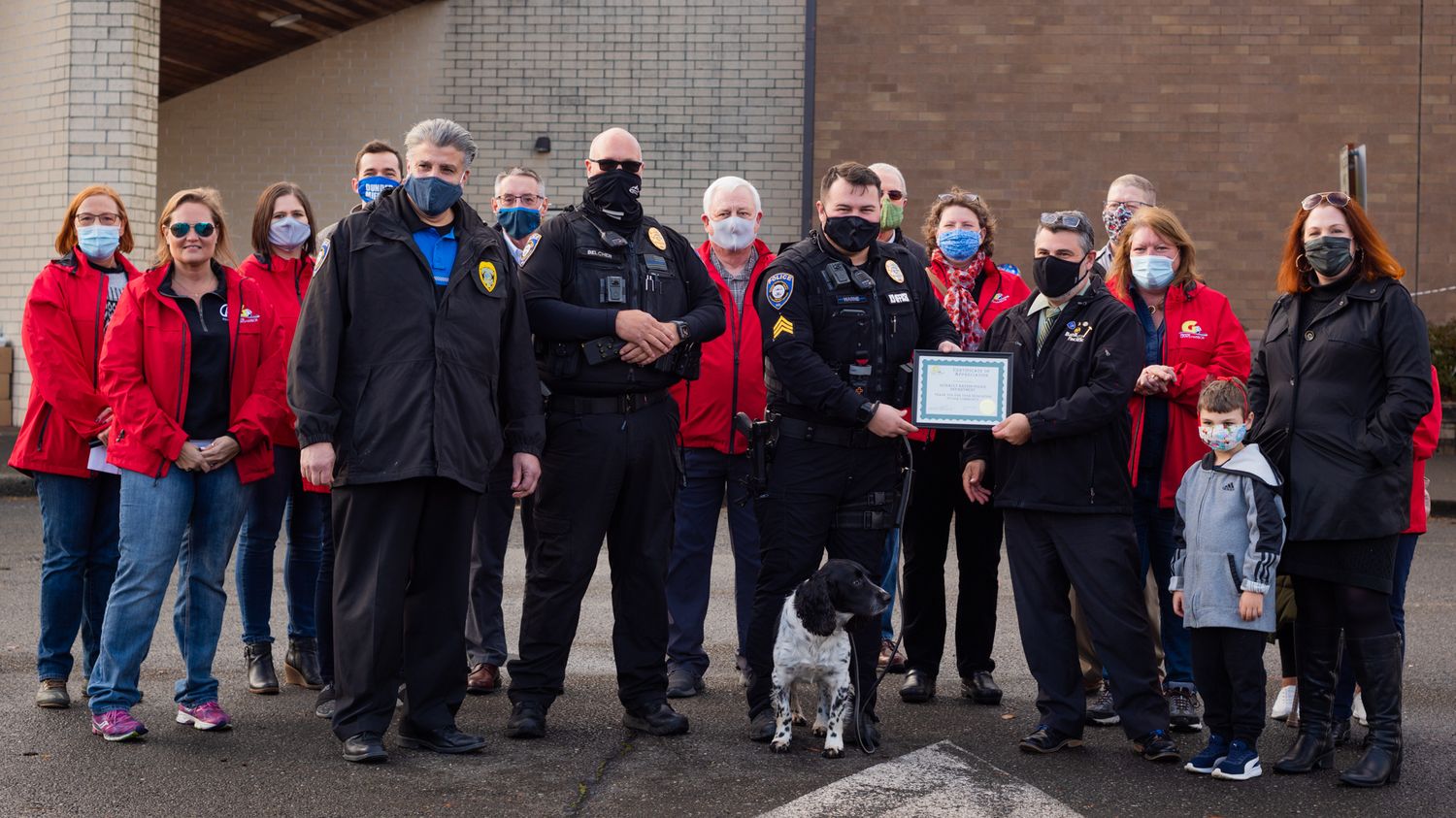 Masked police officers and community members pose with a framed award and a small dog standing in front of them outdoors.