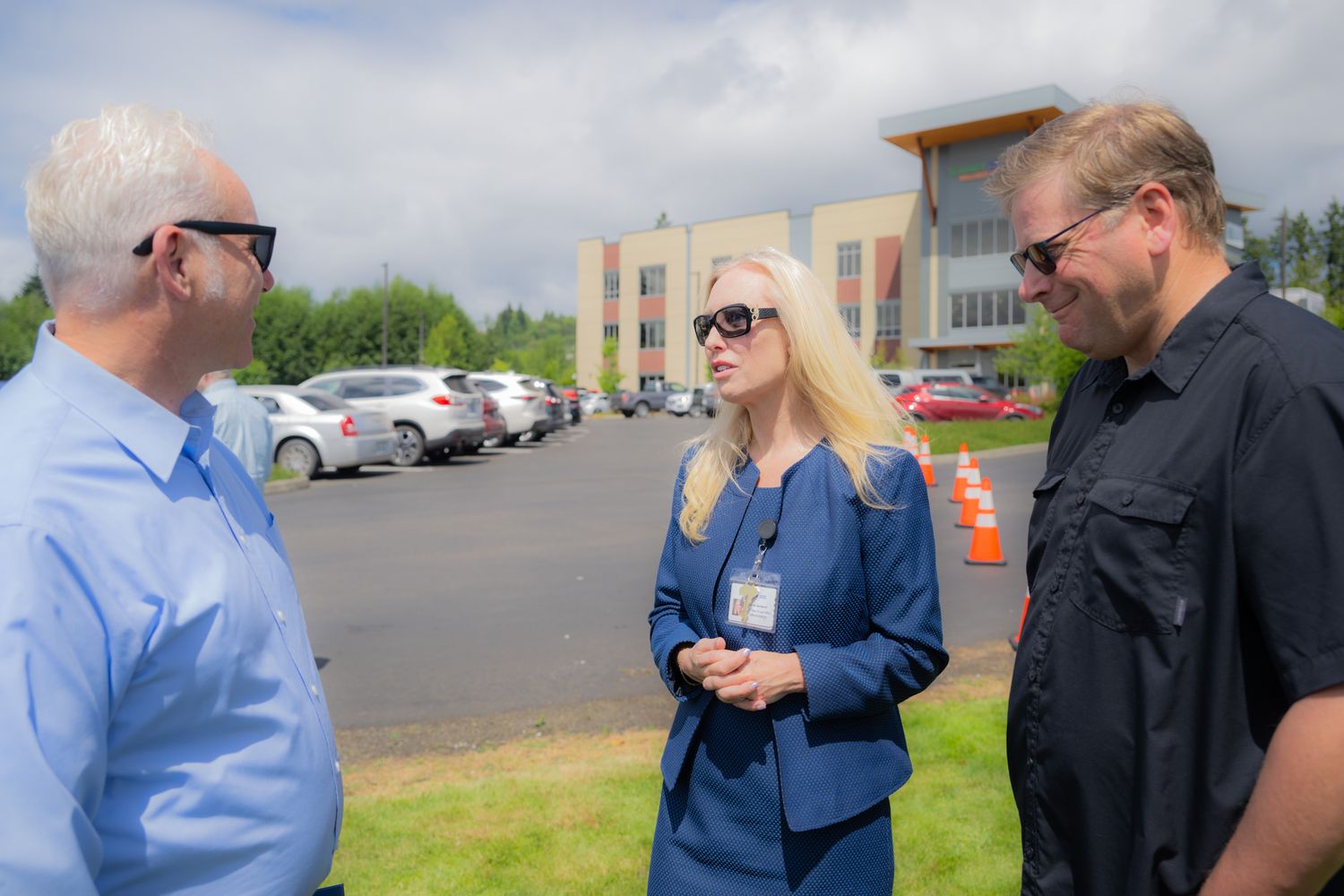 Three people in sunglasses stand outdoors talking in a parking lot, with a blonde woman in a blue blazer and a modern building behind them.