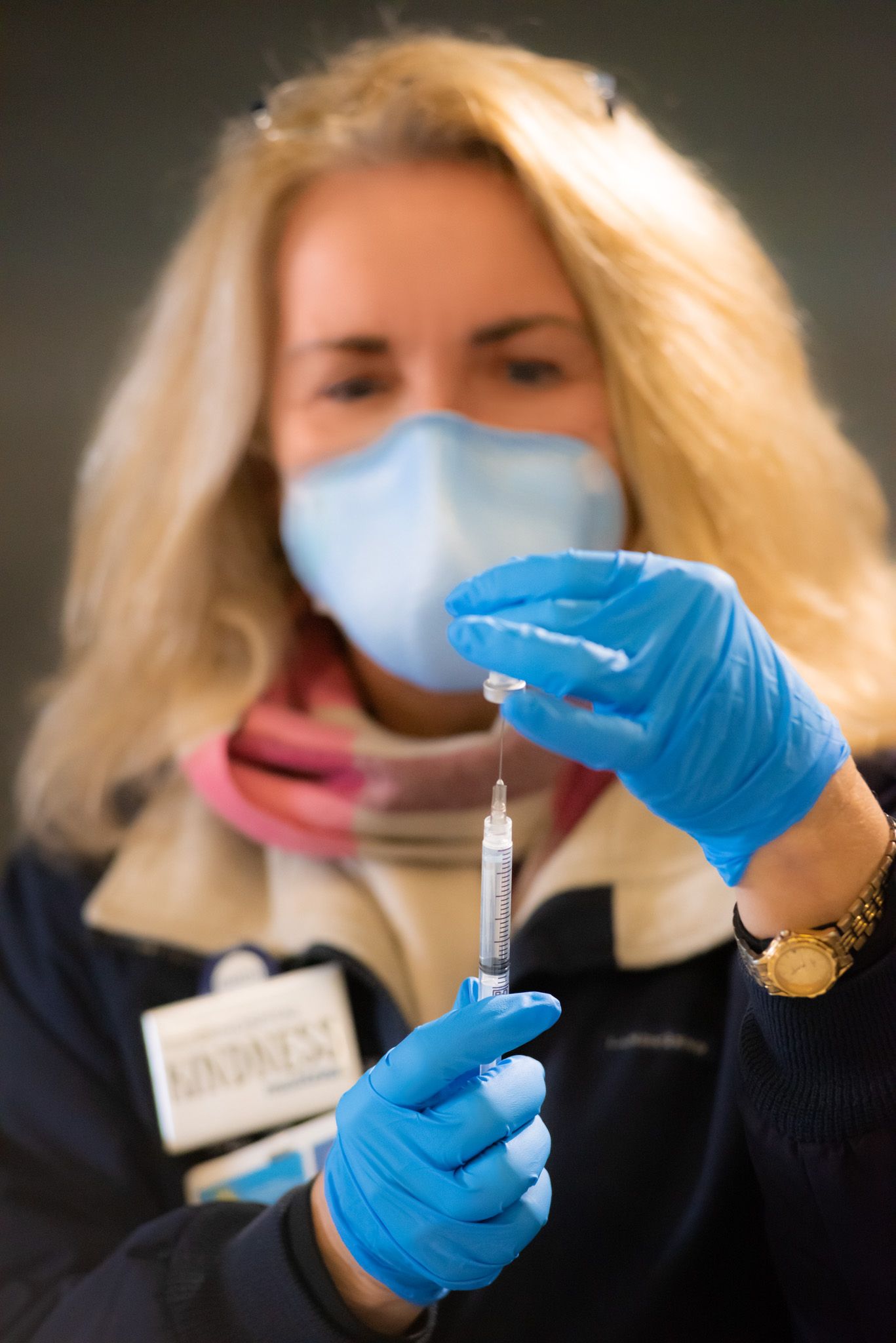 Healthcare worker in a blue face mask and gloves prepares a vaccine dose, holding a syringe up to the light.
