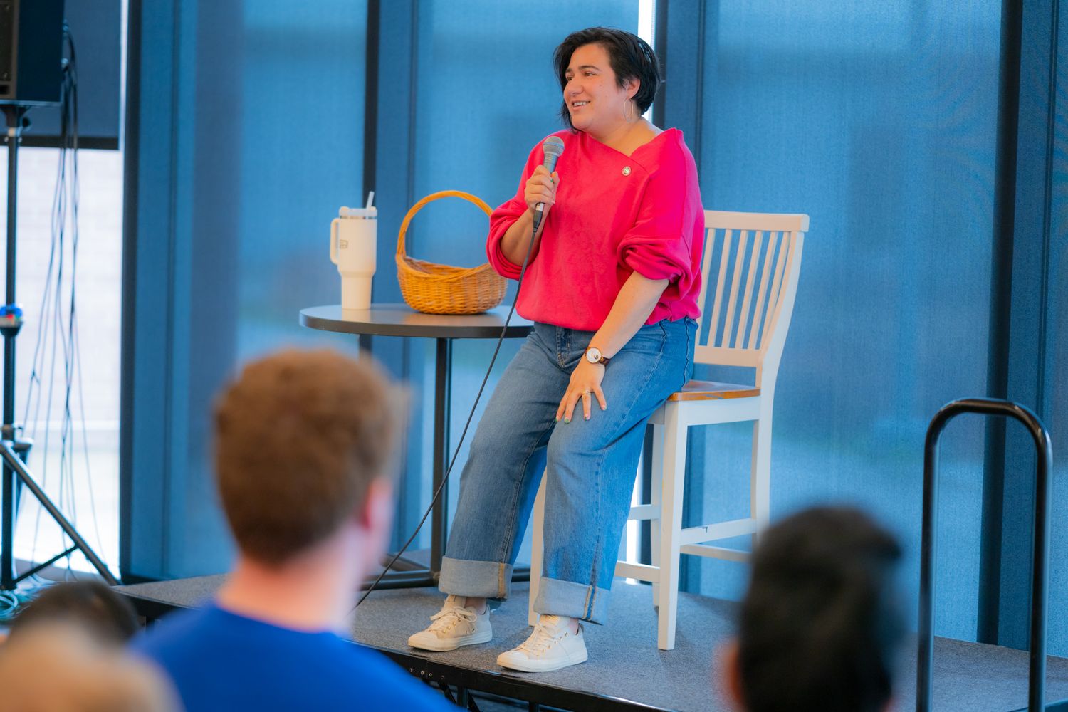 A woman in a pink blouse and jeans sits on a stool holding a microphone while speaking to an audience on a small stage.