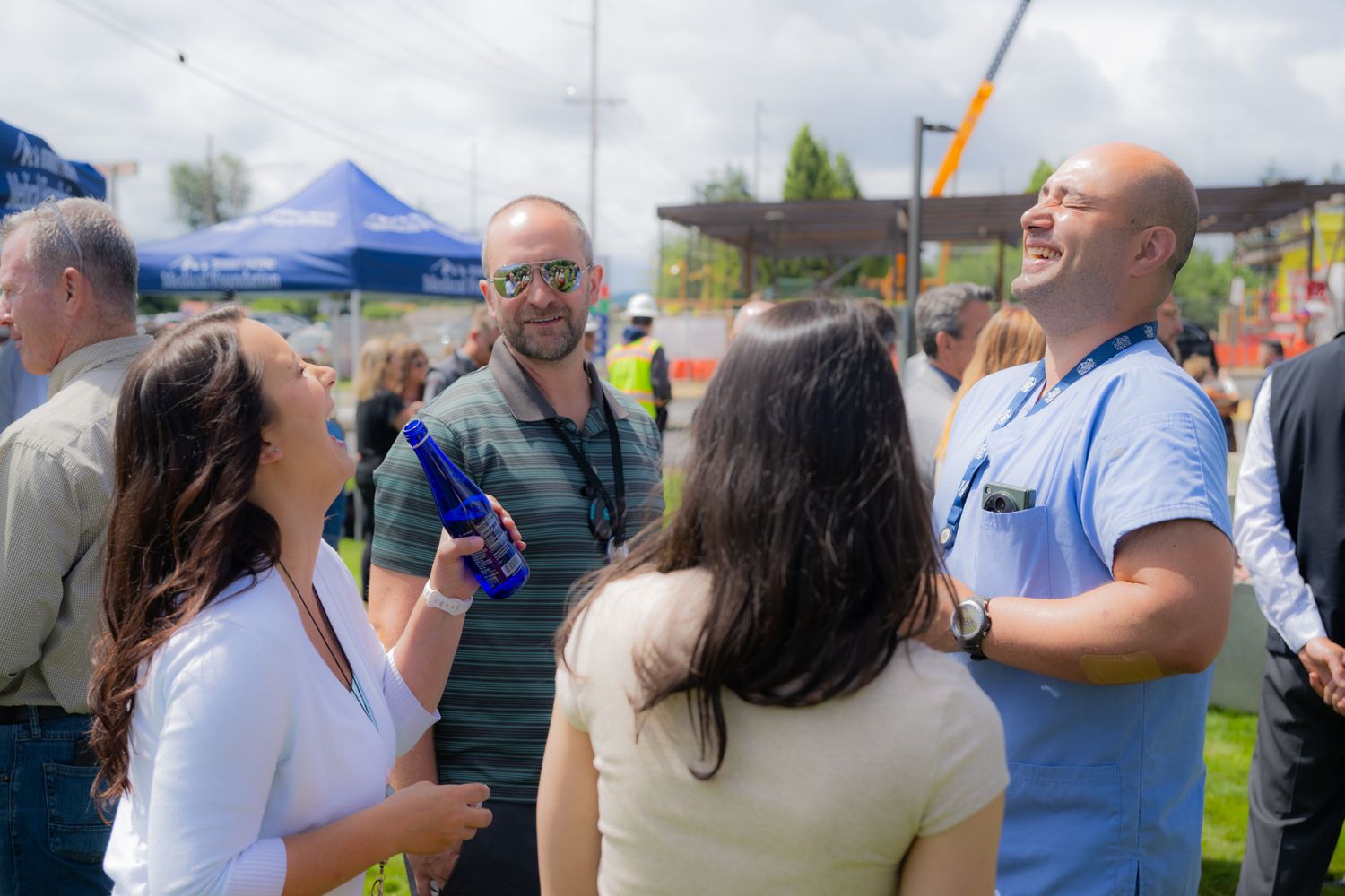 Group of smiling people chatting at an outdoor event with blue canopy tents, one woman holding a blue bottle.