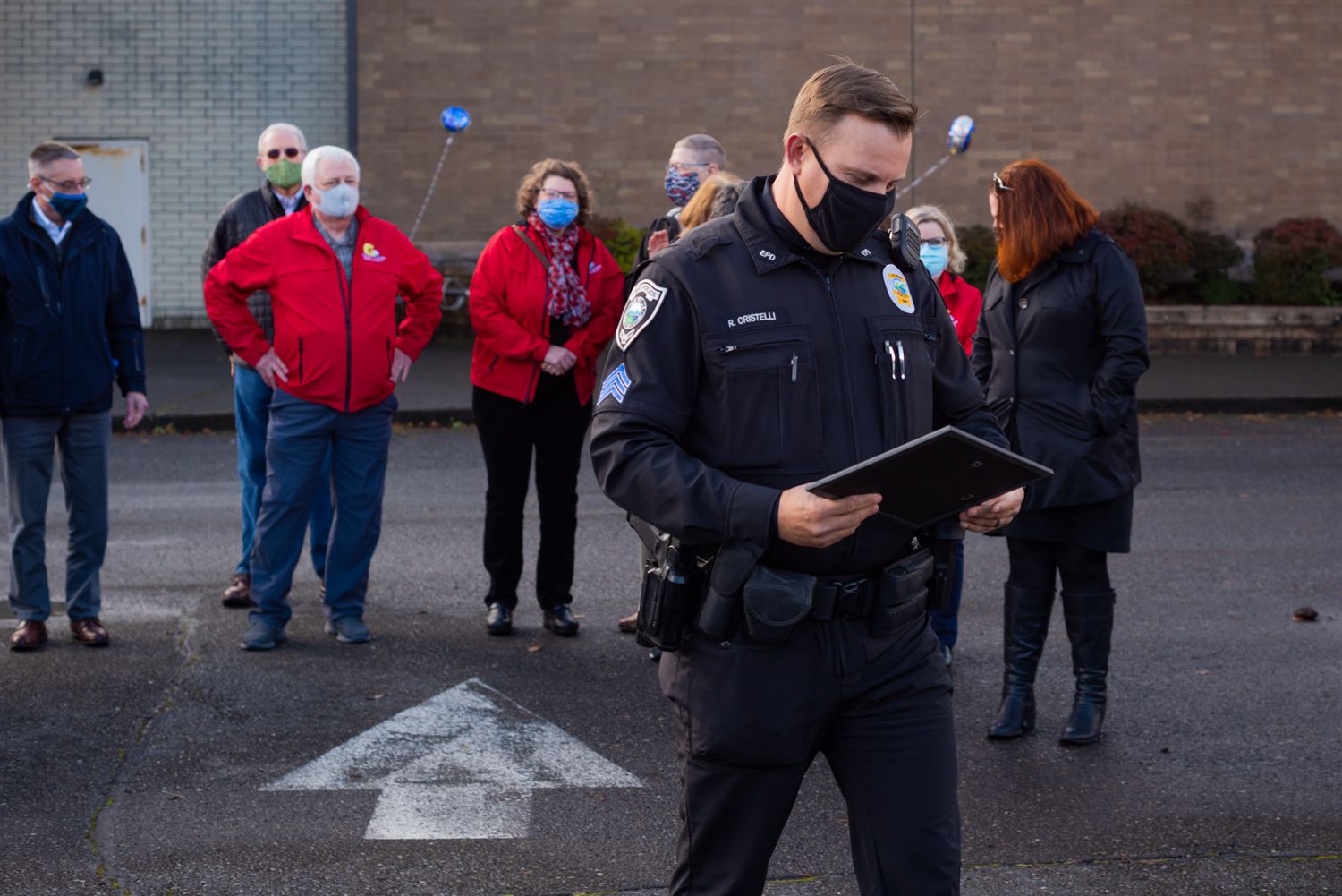 Masked police officer in uniform reads from a folder while a small group of people in face masks stands outdoors on a paved lot behind him.