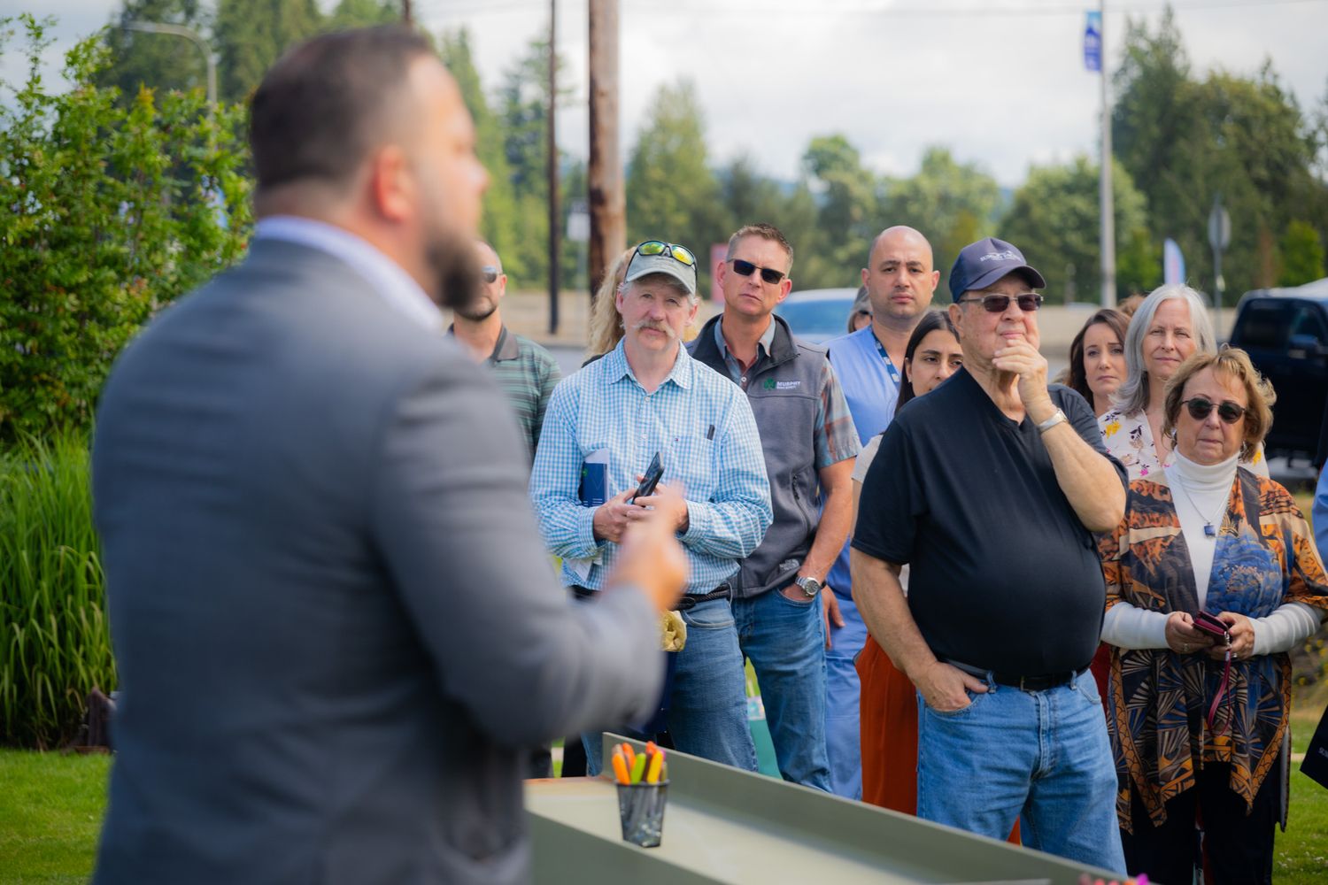 A man in a gray suit speaks to a group of attentive onlookers standing outdoors on a grassy lawn surrounded by trees.