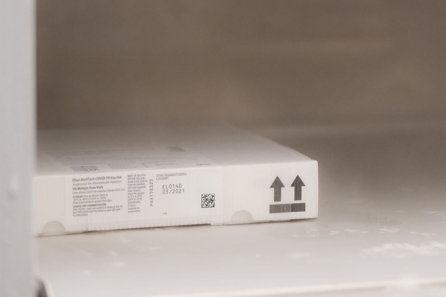 White cardboard shipping box with printed label, barcode, and two upward arrows sitting on a white shelf.