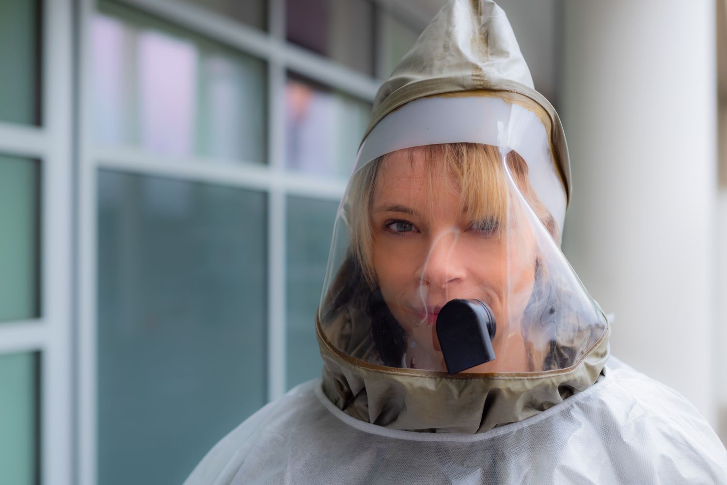 Woman wearing a protective hood respirator with a clear face shield and gray hooded suit standing indoors near large windows.