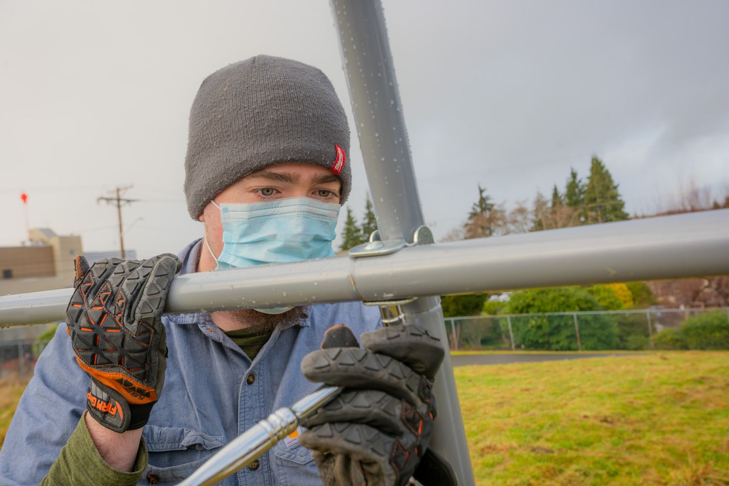 Worker in a gray beanie, blue face mask and work gloves assembling a metal pipe frame outdoors on a grassy field.