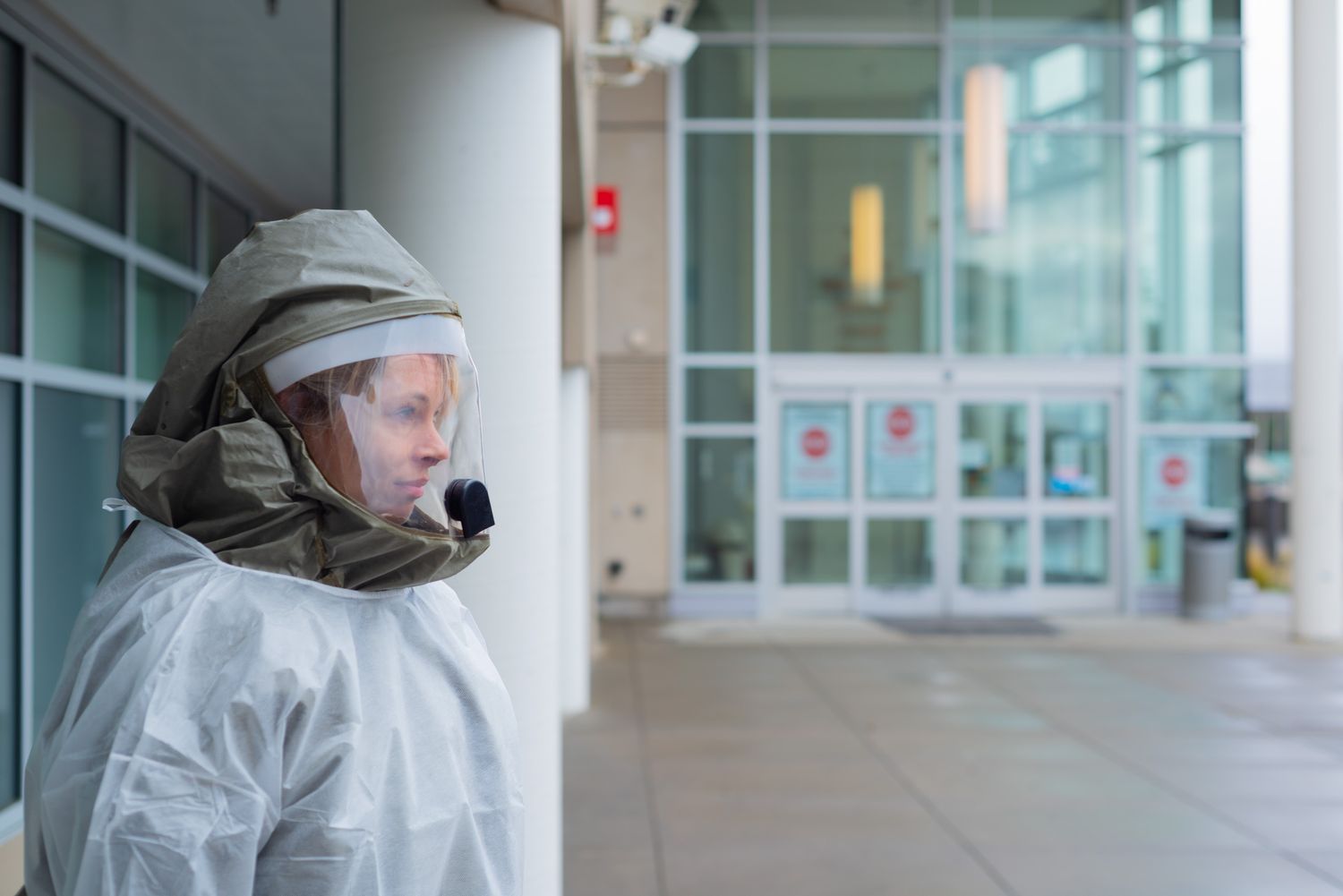 Healthcare worker in a protective gown and hooded face shield stands outside a hospital entrance with glass doors.