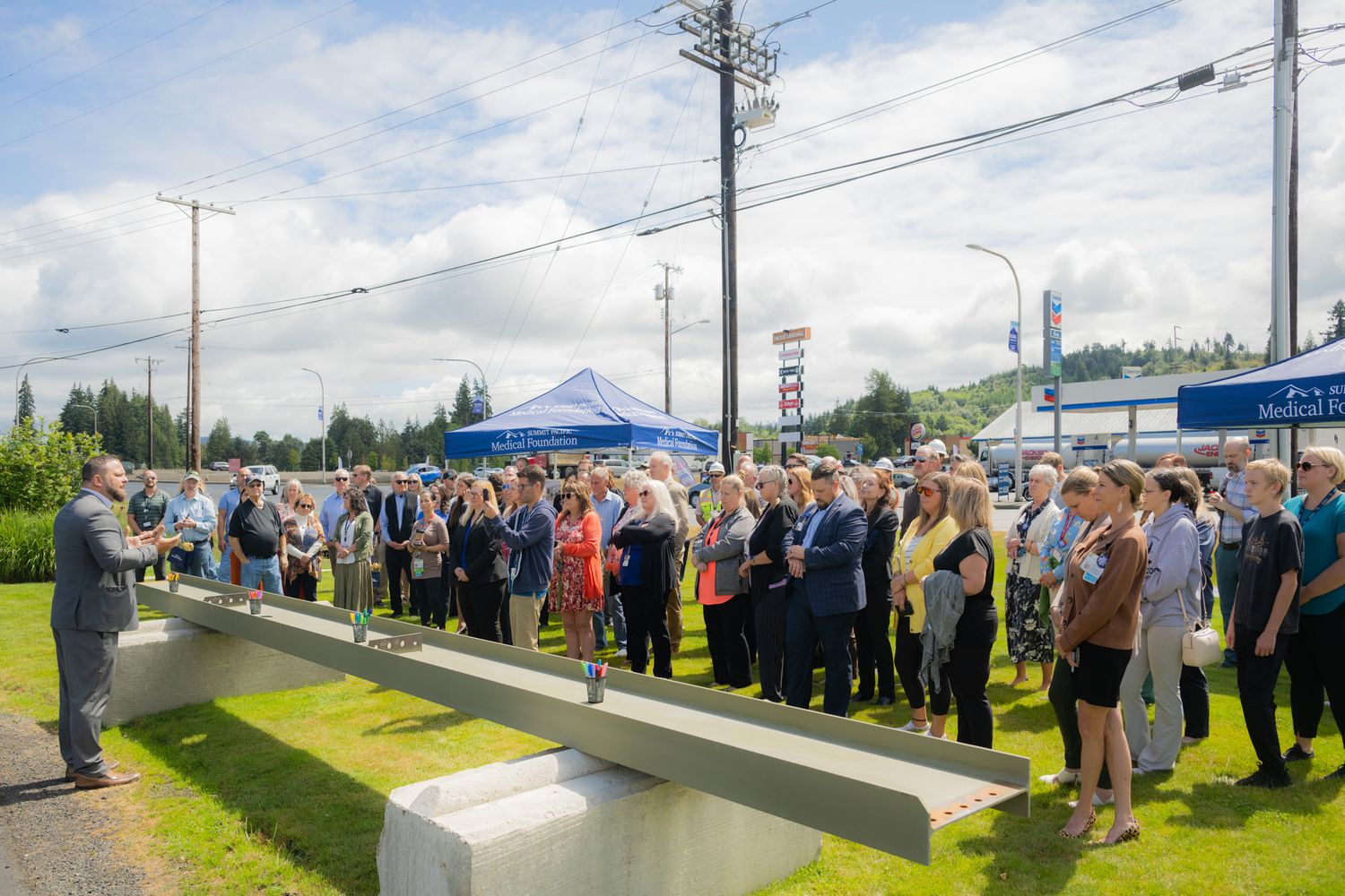Crowd of people gathers outdoors on a grassy lawn near a concrete beam structure, with a blue tent and utility poles in the background.