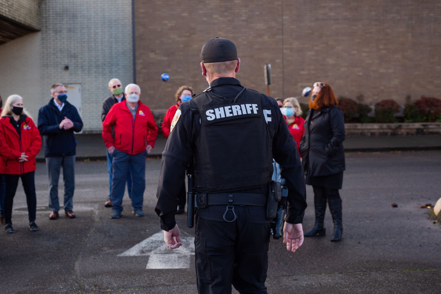 A sheriff's deputy in a black uniform walks toward a small group of masked people gathered outside a brick building.