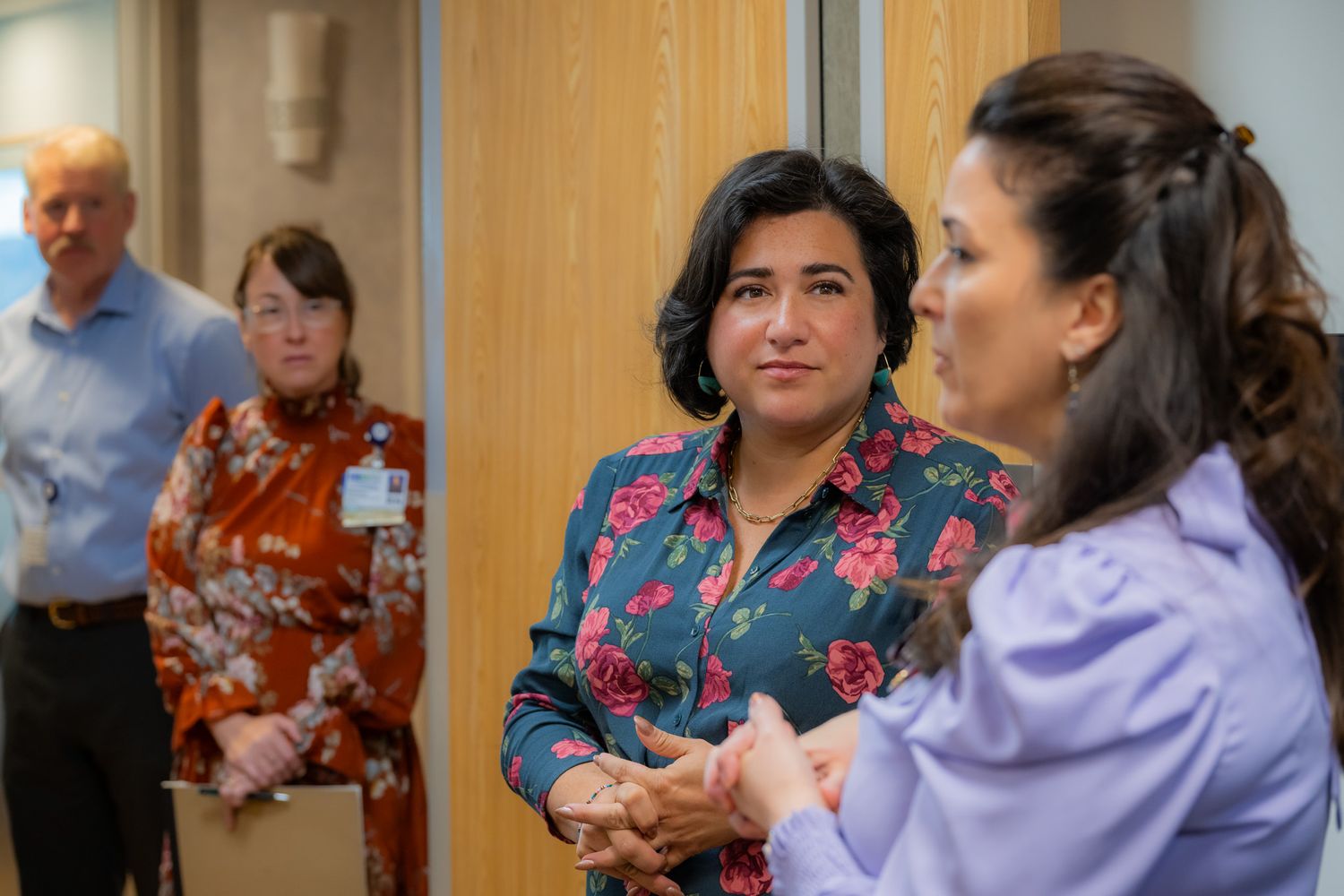 Two women in colorful blouses clasp hands and talk in a professional office while a man and a woman with a name badge stand behind them.