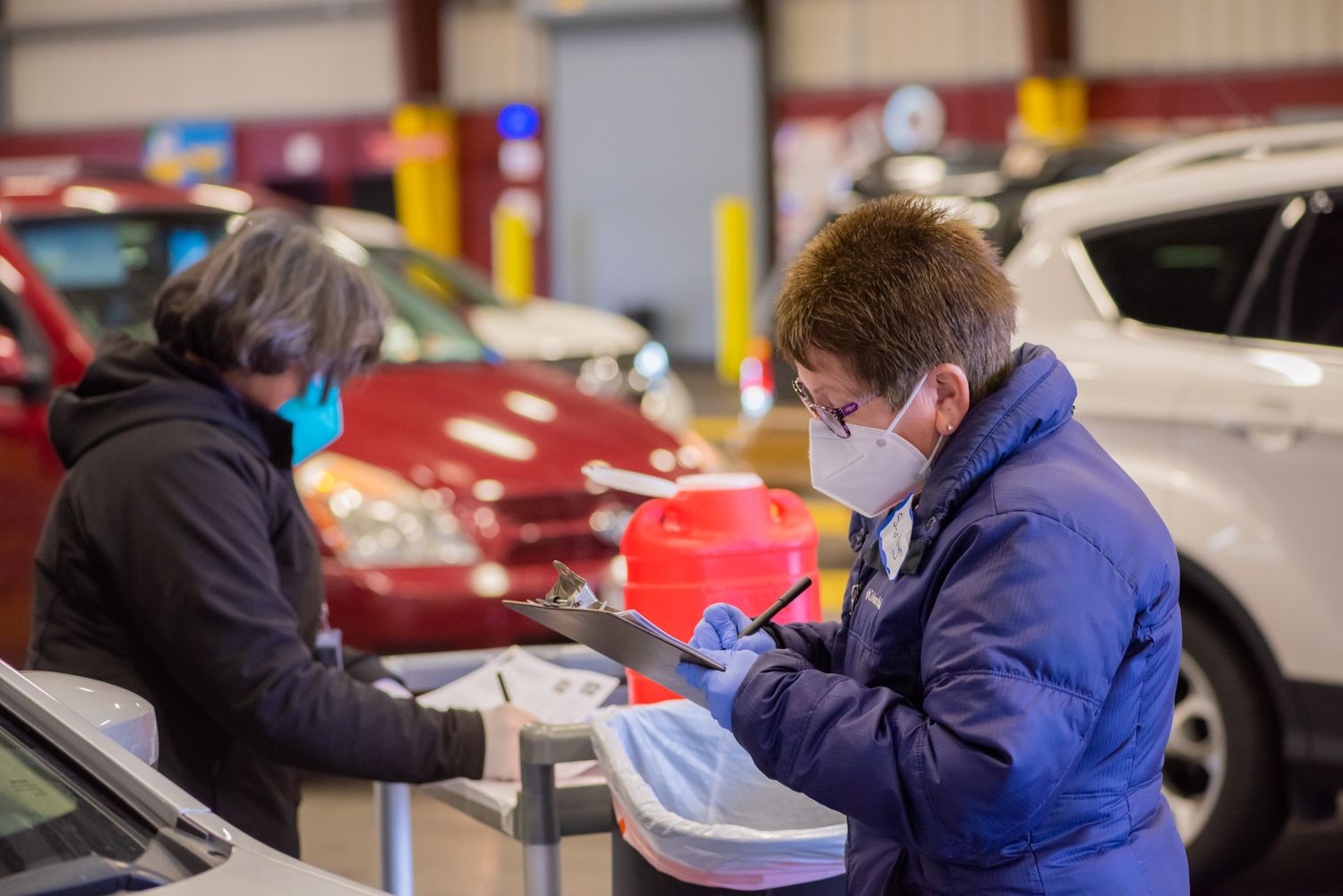 Two masked workers in jackets and gloves review a clipboard at a vehicle inspection station inside a large indoor facility.