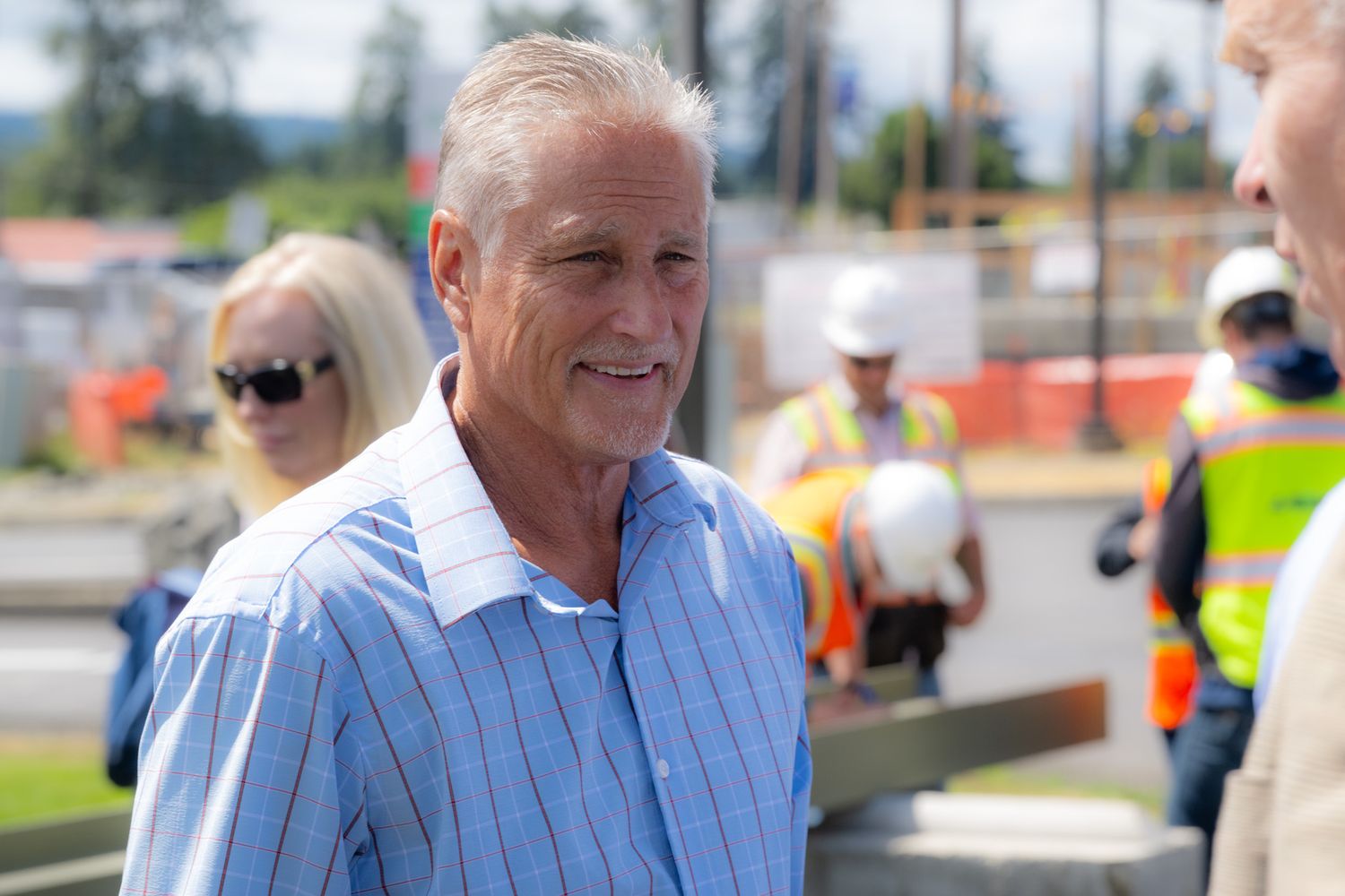 Smiling older man with gray hair in a light blue striped shirt stands at an outdoor construction site with workers in hard hats behind him.