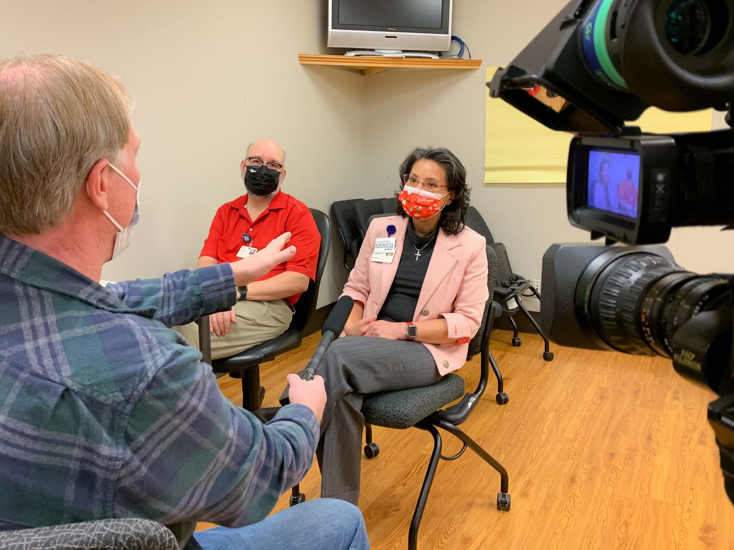 Two masked people sit in chairs being interviewed by a man in a plaid shirt while a video camera records in a bright room.