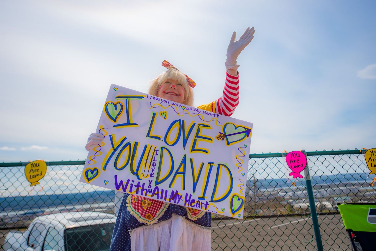 Person in a colorful clown costume waves while holding a handmade sign reading I love you David with all my heart beside a fence.
