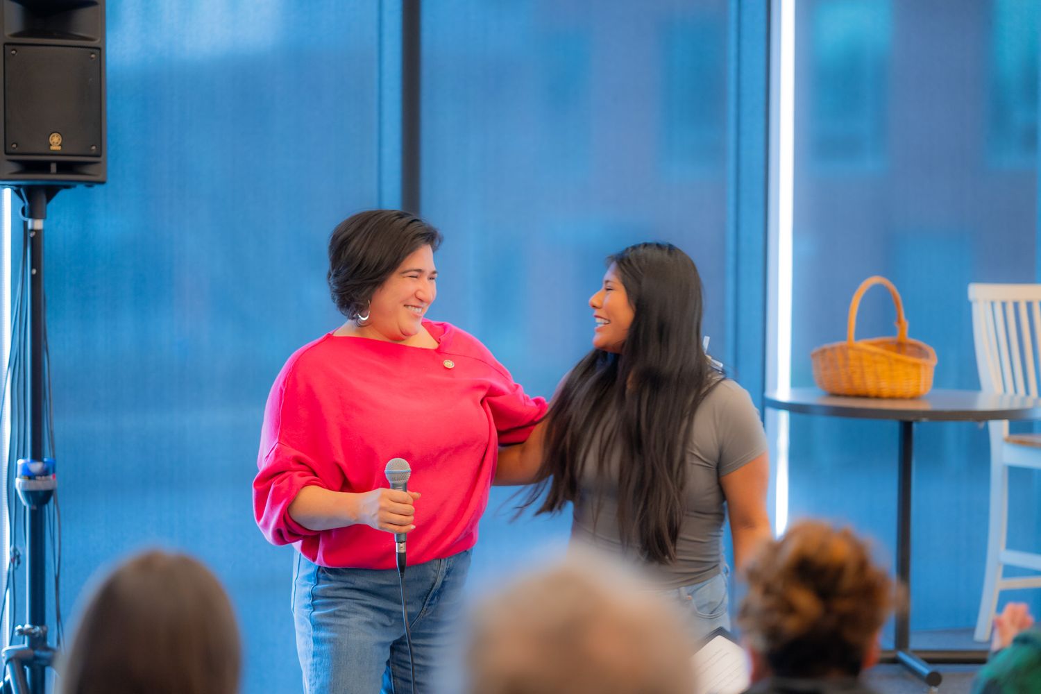 Two smiling women stand side by side on a stage, one holding a microphone, as audience members watch near large windows.