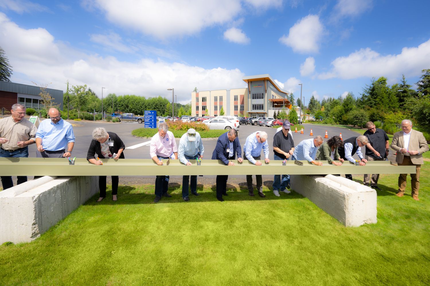 Group of people in business casual attire stand in a row signing a long beam resting on concrete blocks on a green lawn with a building behi