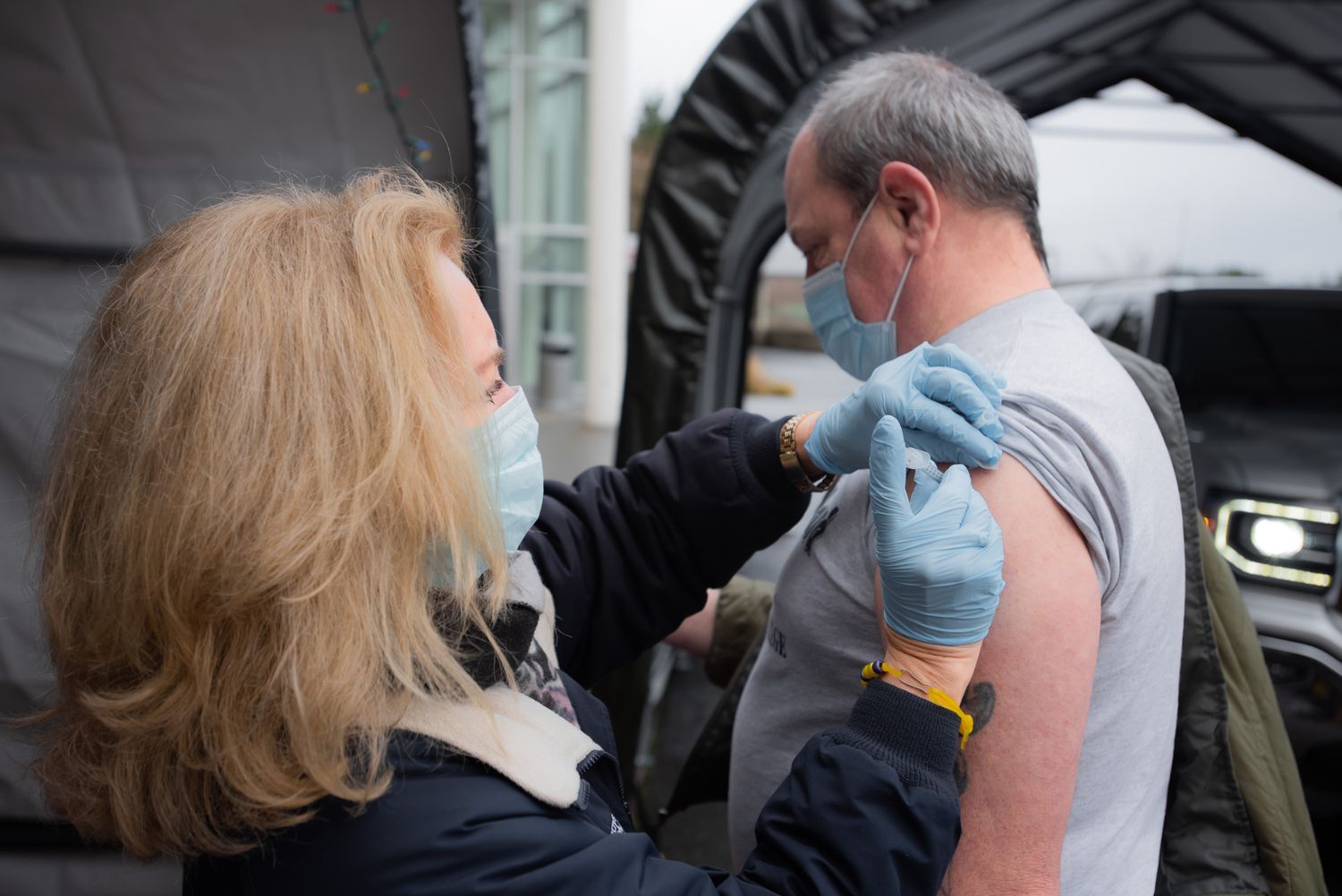 A masked healthcare worker in blue gloves gives a vaccine injection to the upper arm of a masked man at an outdoor drive-through clinic.