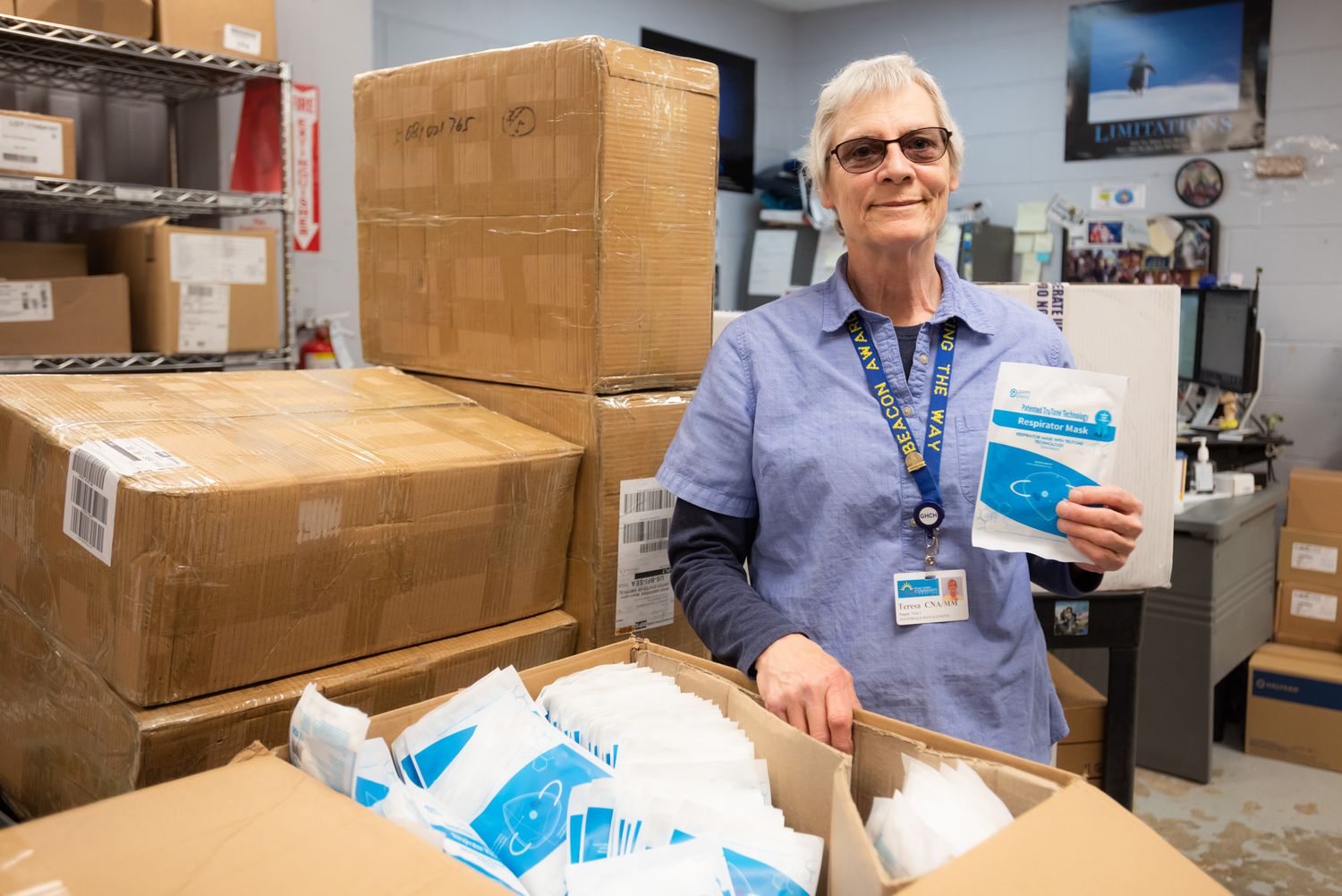 Smiling woman in a blue uniform and lanyard holds a packaged face mask over an open box of masks, with stacked cardboard boxes behind her.