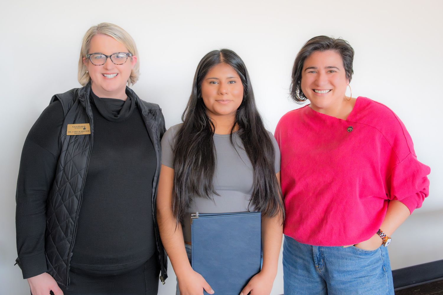 Three smiling women stand side by side indoors, one in a black vest with a name tag, one holding a folder, and one in a bright pink sweater.